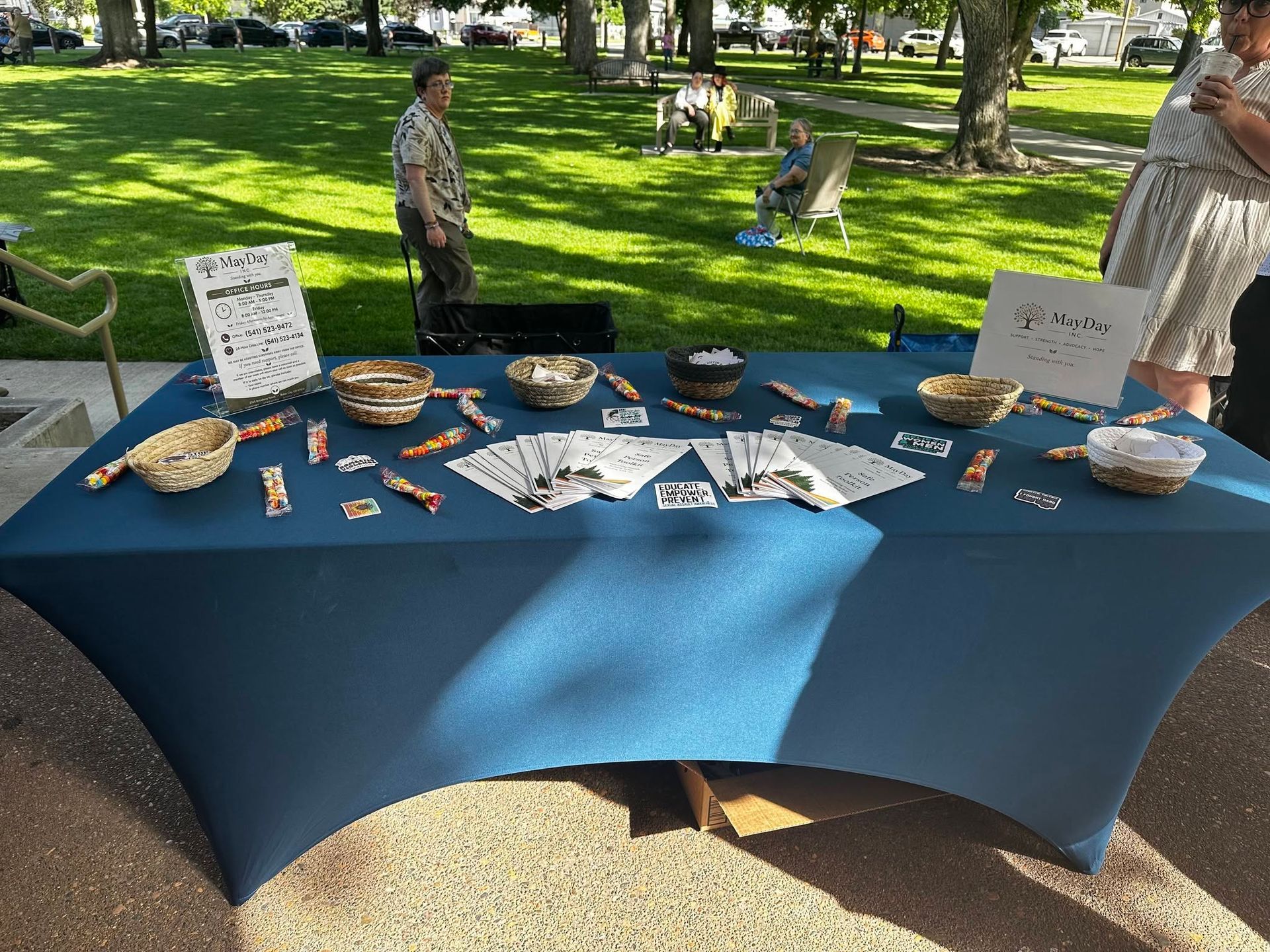 Outdoor park table display with flyers, bowls, and small items on a blue cloth under trees