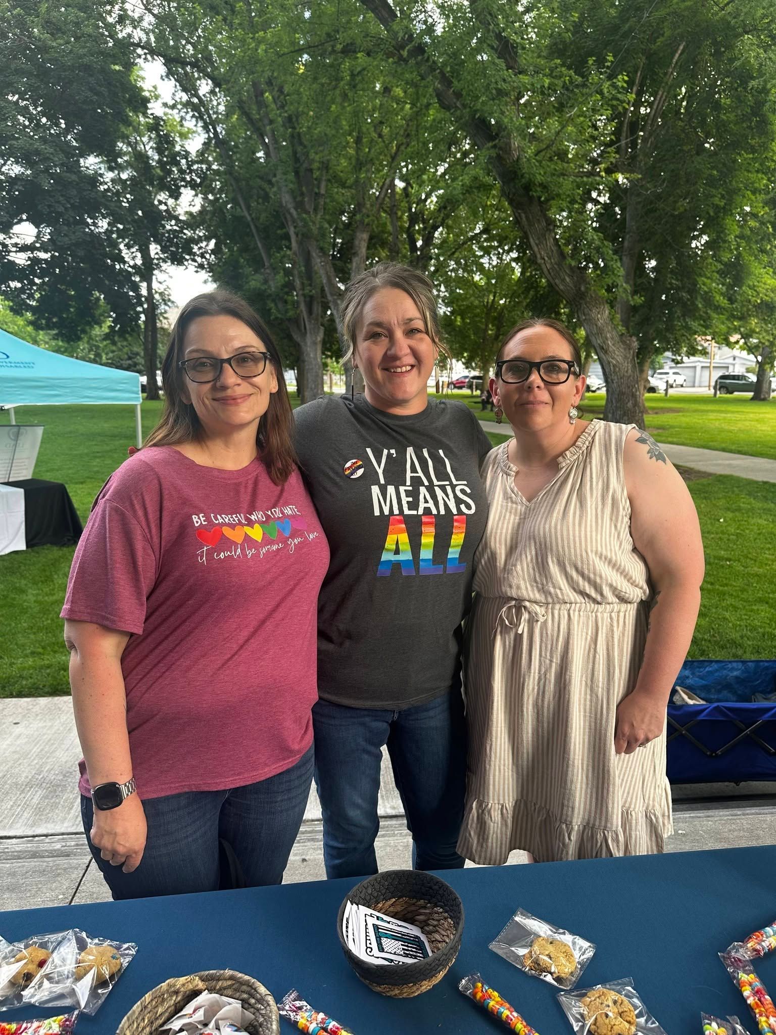 Three people standing behind a table outdoors at a community event, smiling in front of trees.