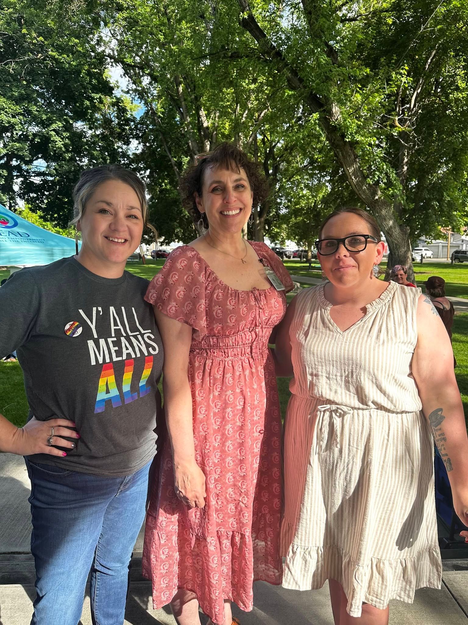 Three women standing outdoors under a leafy tree, smiling in casual summer clothes.