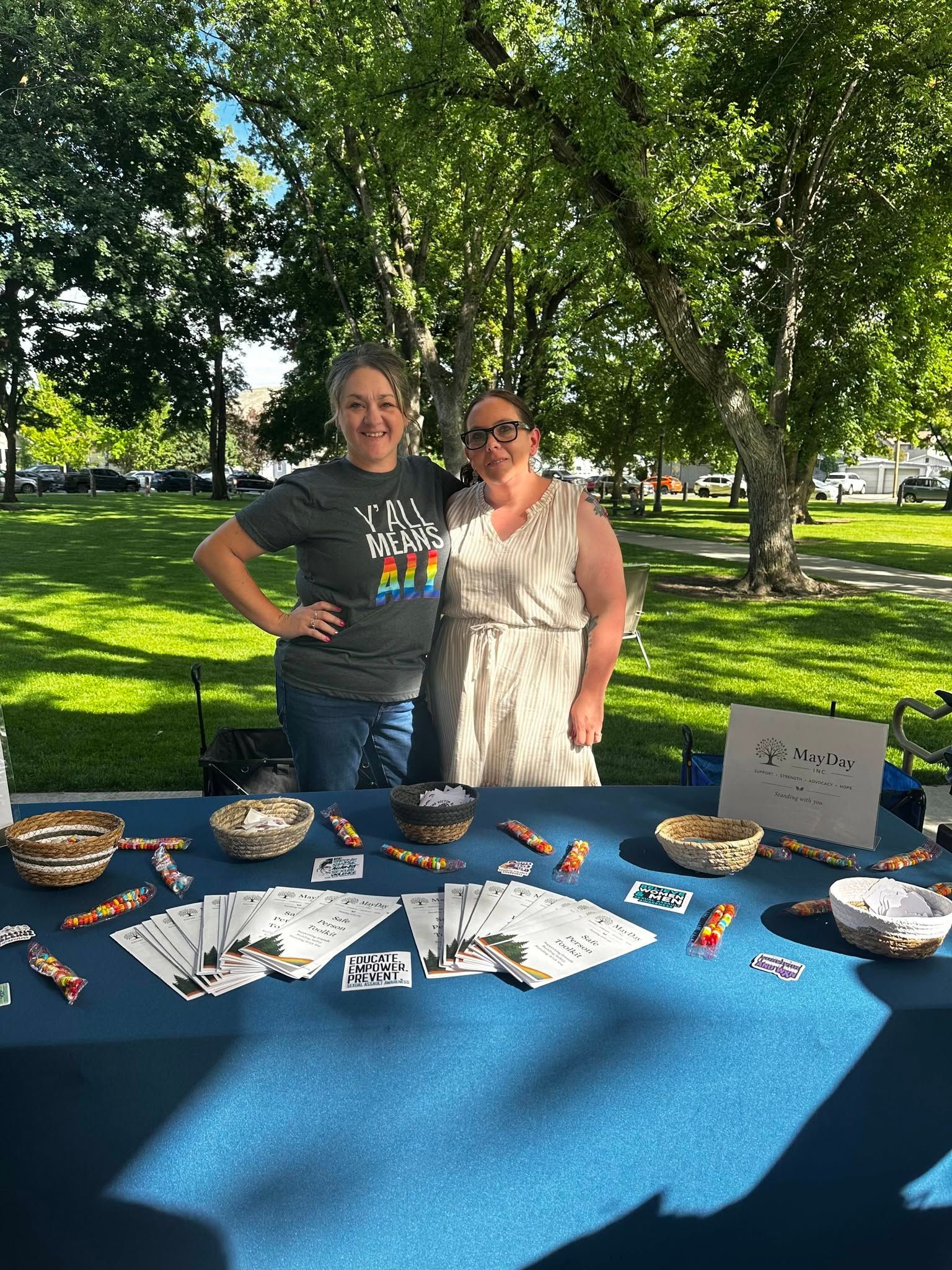 Two people standing behind a blue table at an outdoor park booth with crafts and signs displayed