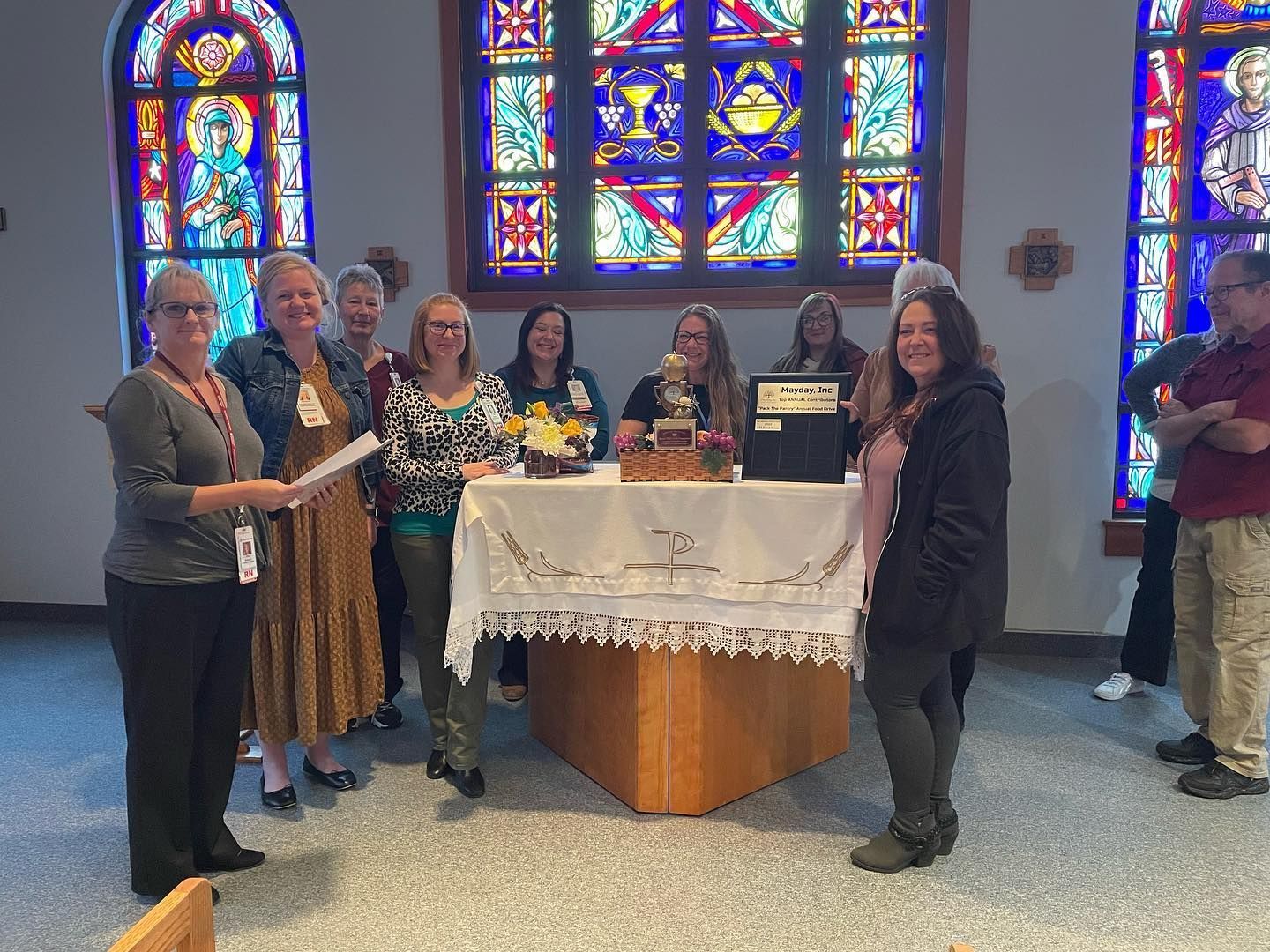 Group posed behind a table with gifts in a church hall, framed by colorful stained-glass windows.