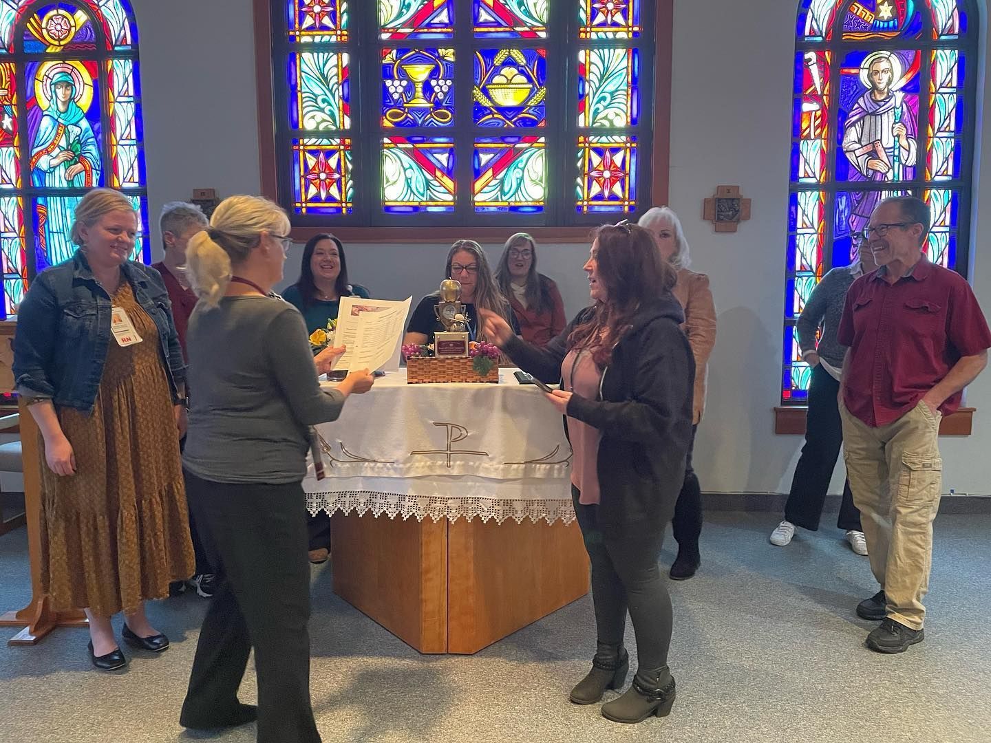 People gathered around a lace-covered table in a stained-glass chapel, chatting and holding items.