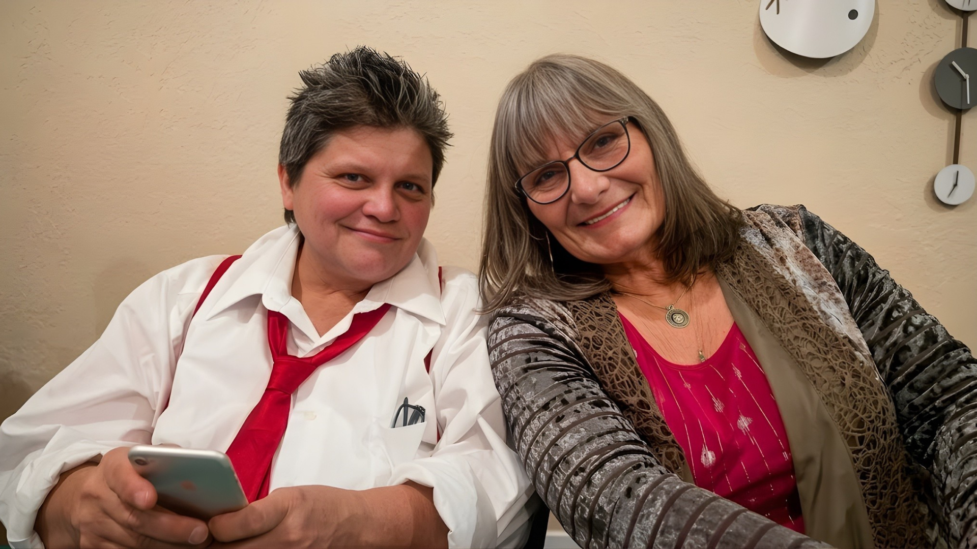 Two smiling people sitting close together, one holding a phone, against a wall with clocks and shelves