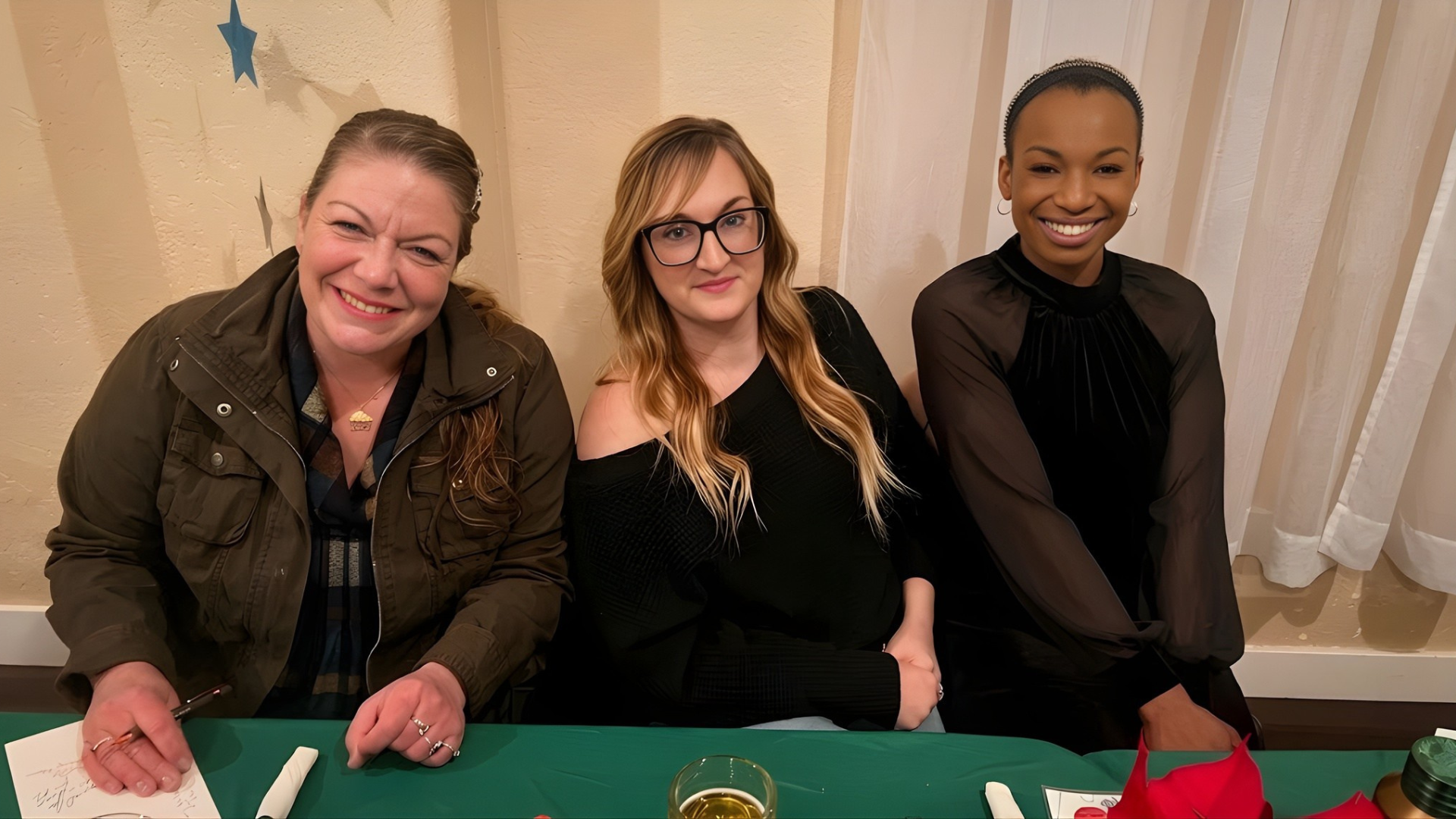 Three people smiling at a table with a green tablecloth in front of beige curtains
