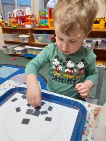 Boy in Mickey Mouse shirt playing with squares on a tray at a table.