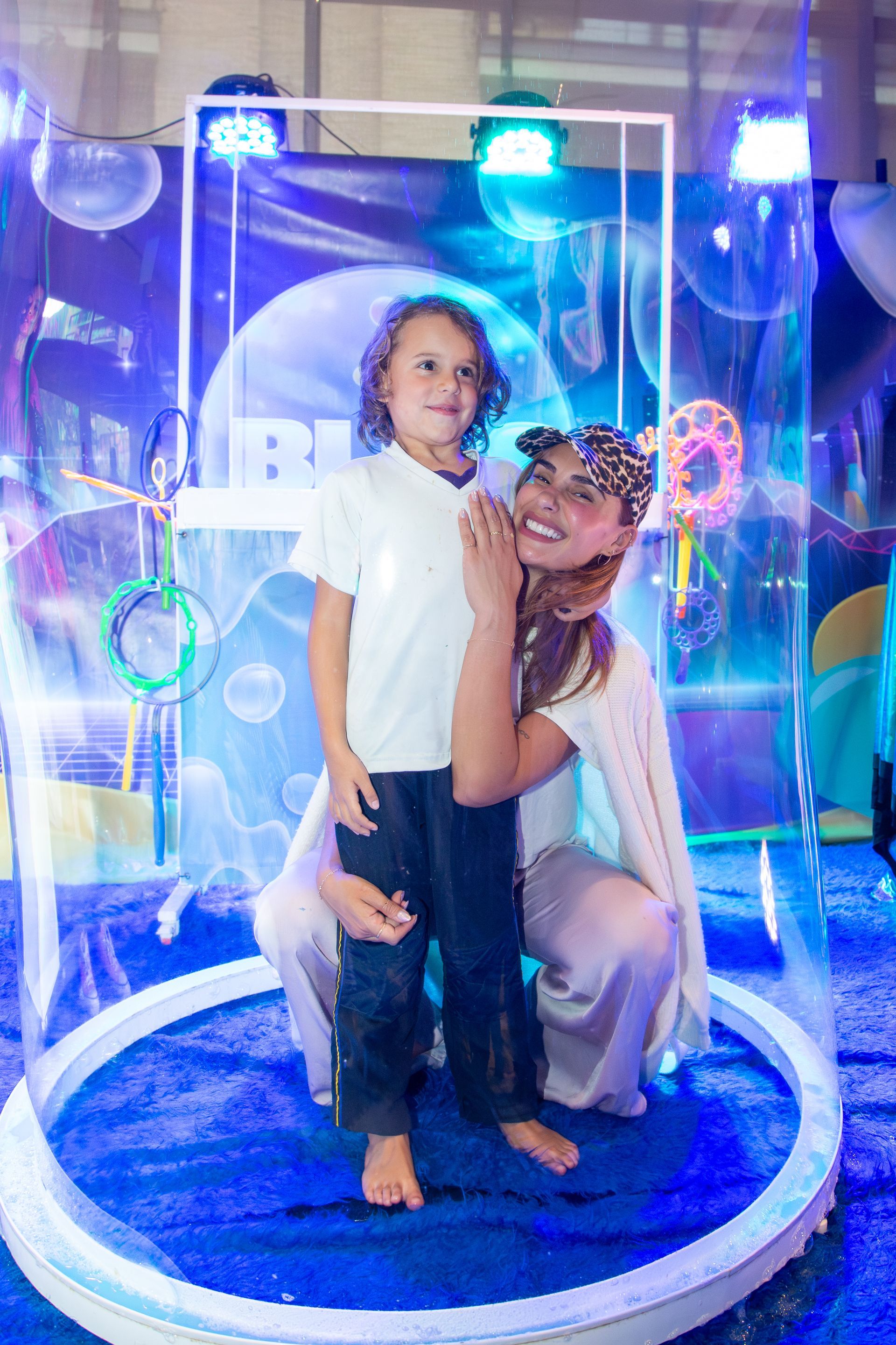 Mujer y niño posando dentro de un marco de burbuja iluminado en un evento con decoraciones azules.