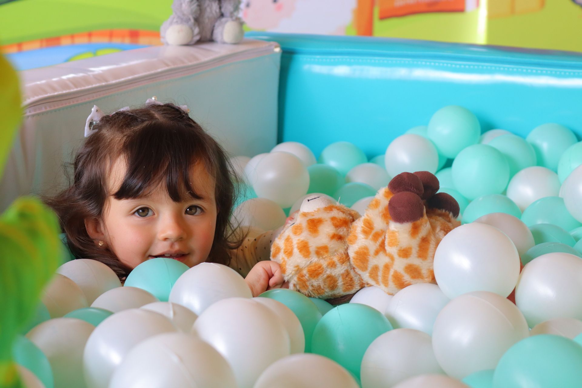 Niña en una piscina de pelotas turquesa con pelotas blancas y verde menta, sosteniendo jirafas de peluche, sonriendo.