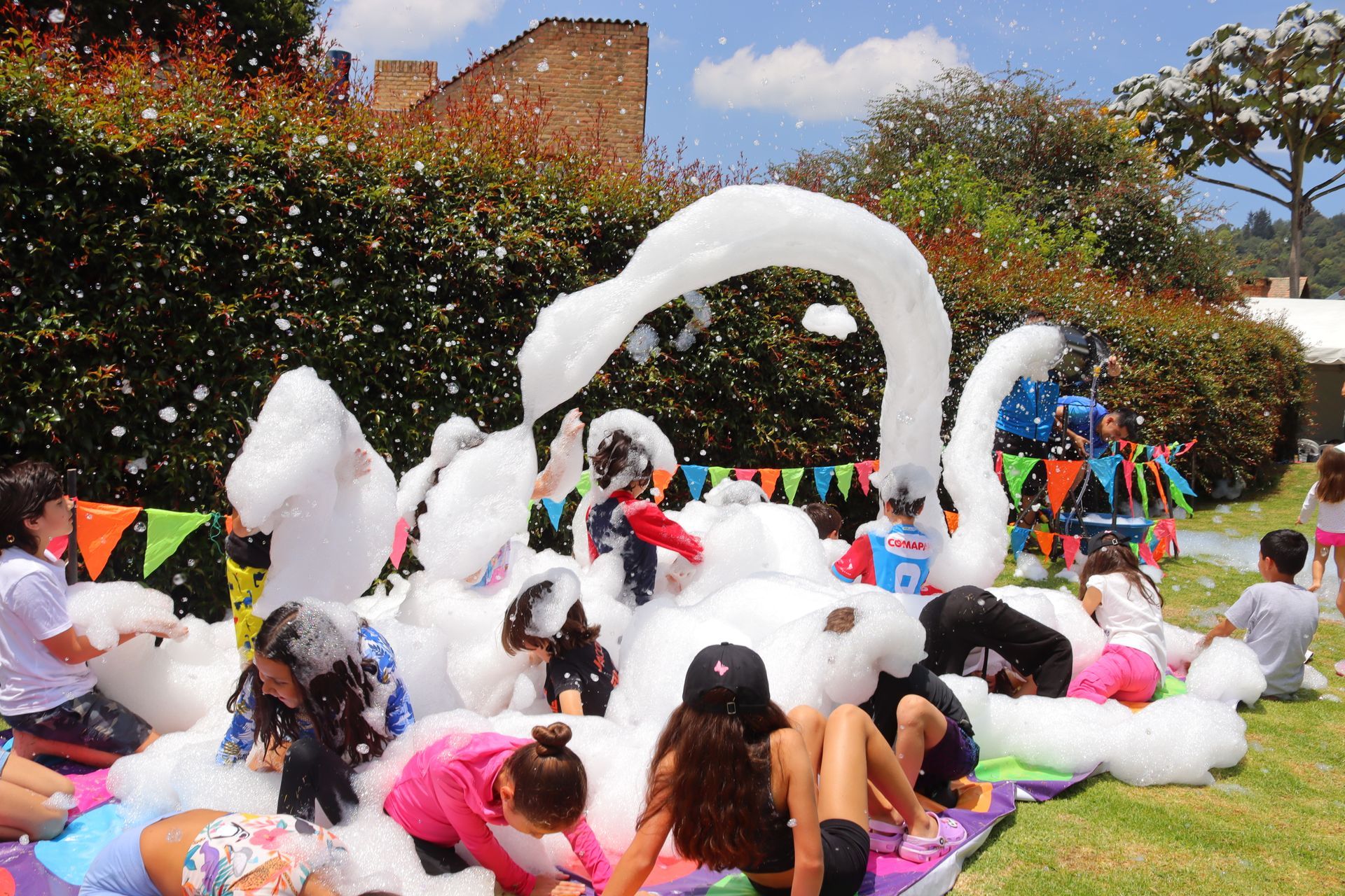 Niños jugando con espuma de una máquina al aire libre, con banderas de colores y árboles.