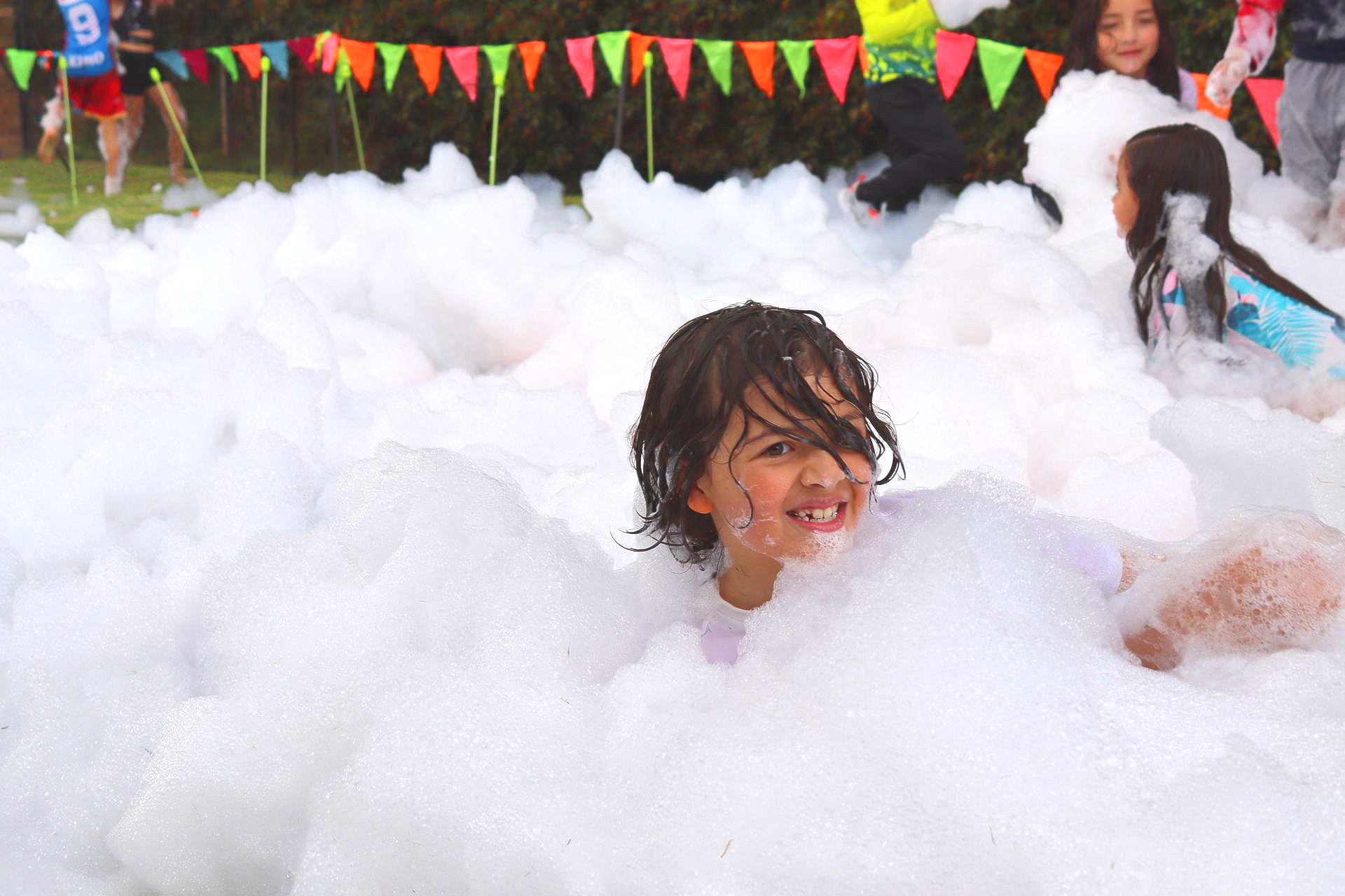 Niño sonriendo en una piscina espumosa en un evento al aire libre, rodeado de otros, con banderas de colores en el fondo.
