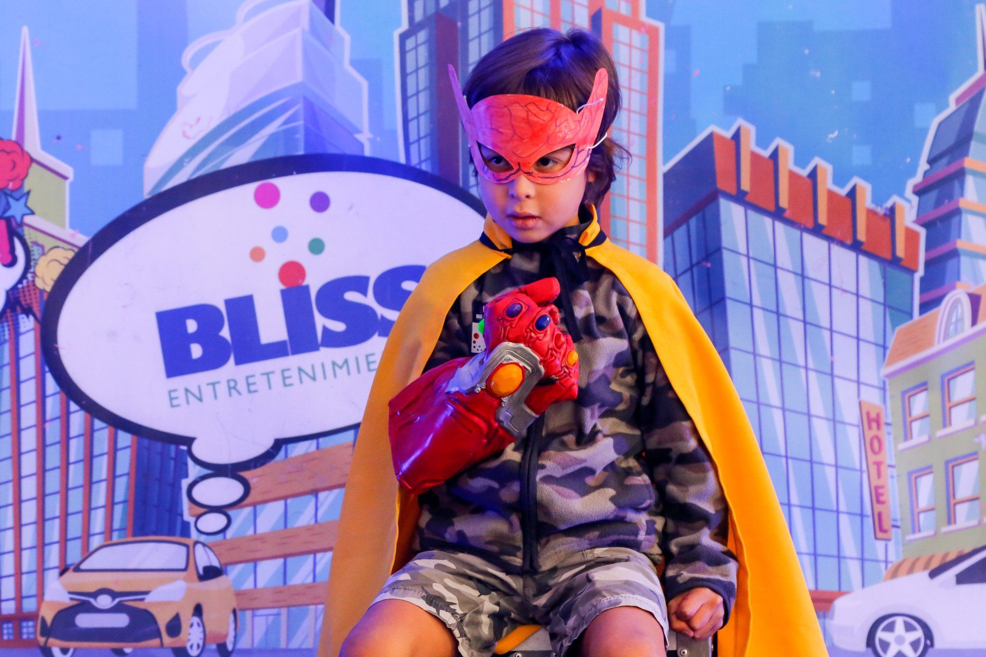 Un niño con un disfraz de superhéroe está sentado frente a un cartel de entretenimiento de Bliss.
