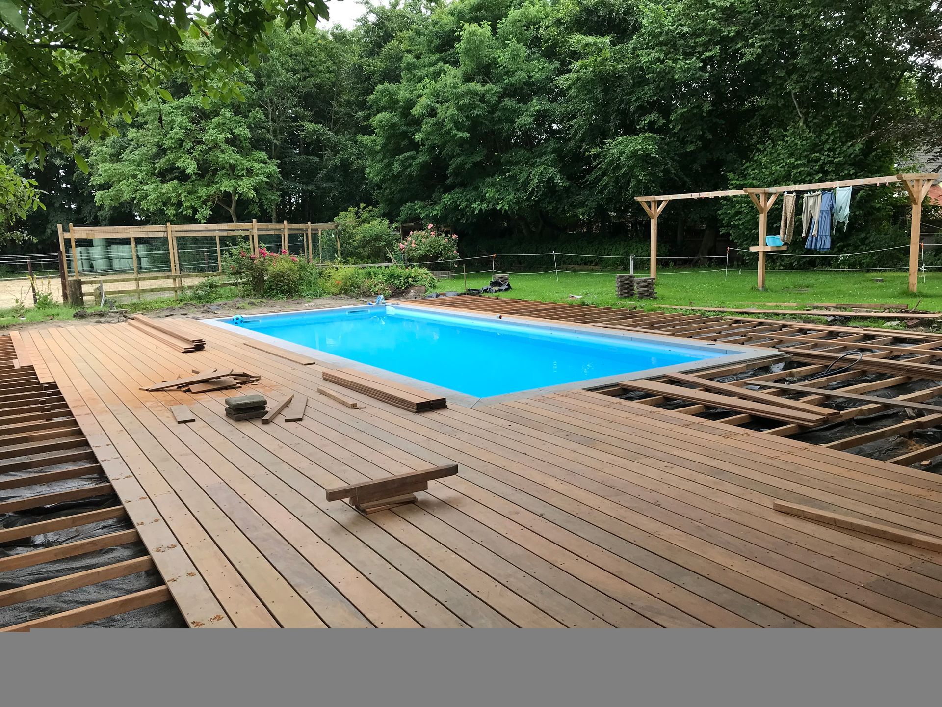 Een rechthoekig blauw zwembad in aanbouw, met houten vlonders eromheen, in een tuinomgeving.
