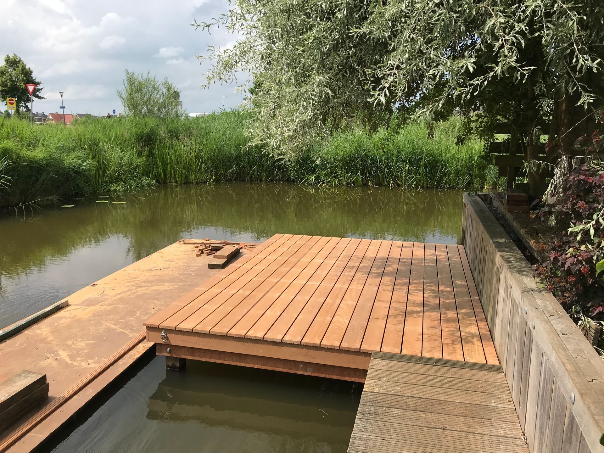 Aan de rand van een rustig, smal kanaal ligt een houten aanlegsteiger, omgeven door weelderig groen riet en loofbomen.