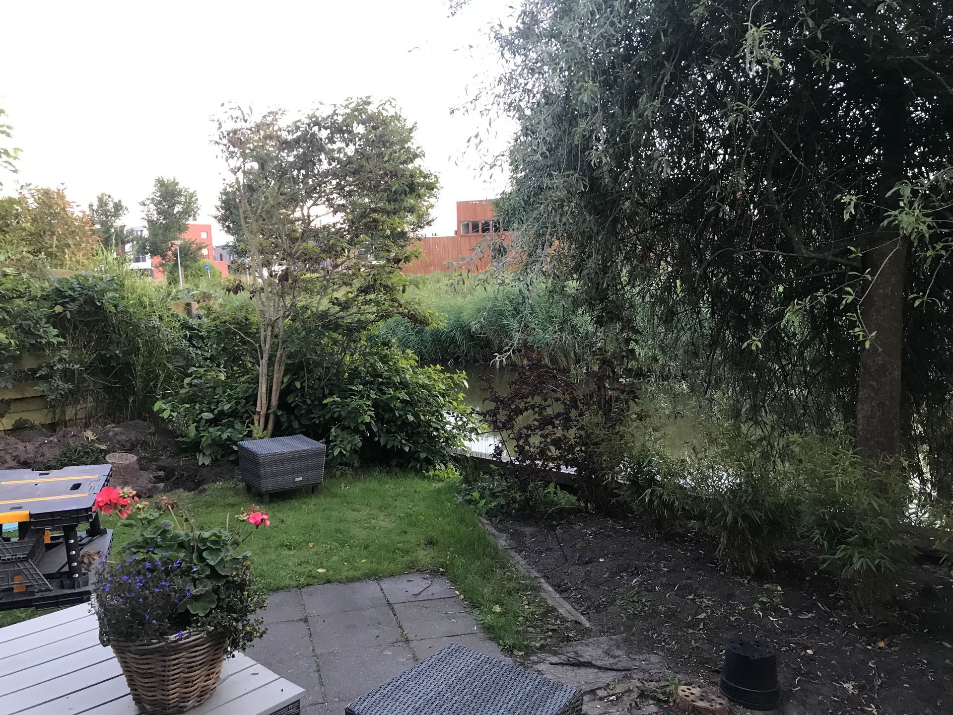 Een achtertuin met uitzicht op een kleine vijver, omgeven door weelderige groene bomen en struiken, met een stenen terras en tuinmeubilair op de voorgrond.