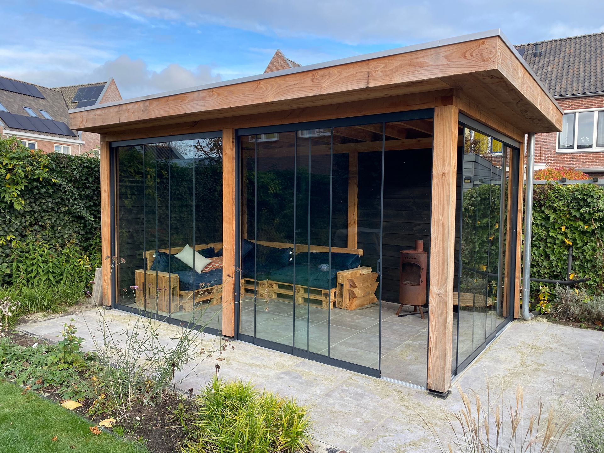 Modern houten tuinpaviljoen met glazen wanden en schuifdeuren, met daarin een bank in palletstijl, geplaatst op een stenen terras.