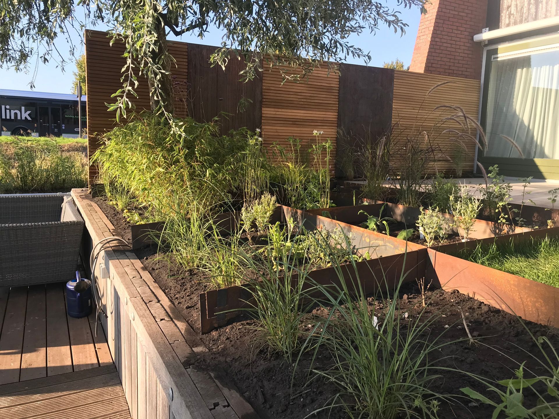 Verhoogde tuinbedden met donkere aarde, groene planten en een rustiek metalen scherm, geplaatst tegen een bakstenen gebouw en een houten terras.