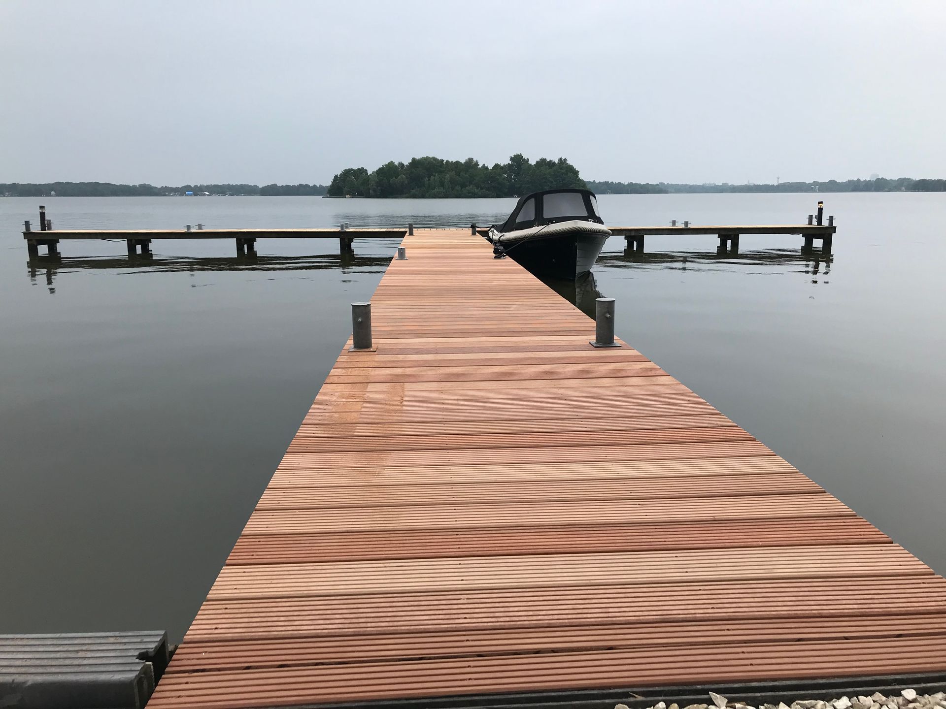 Een houten steiger loopt een rustig meer in, richting een klein eilandje, met een boot die aan het einde van de loopbrug ligt aangemeerd.