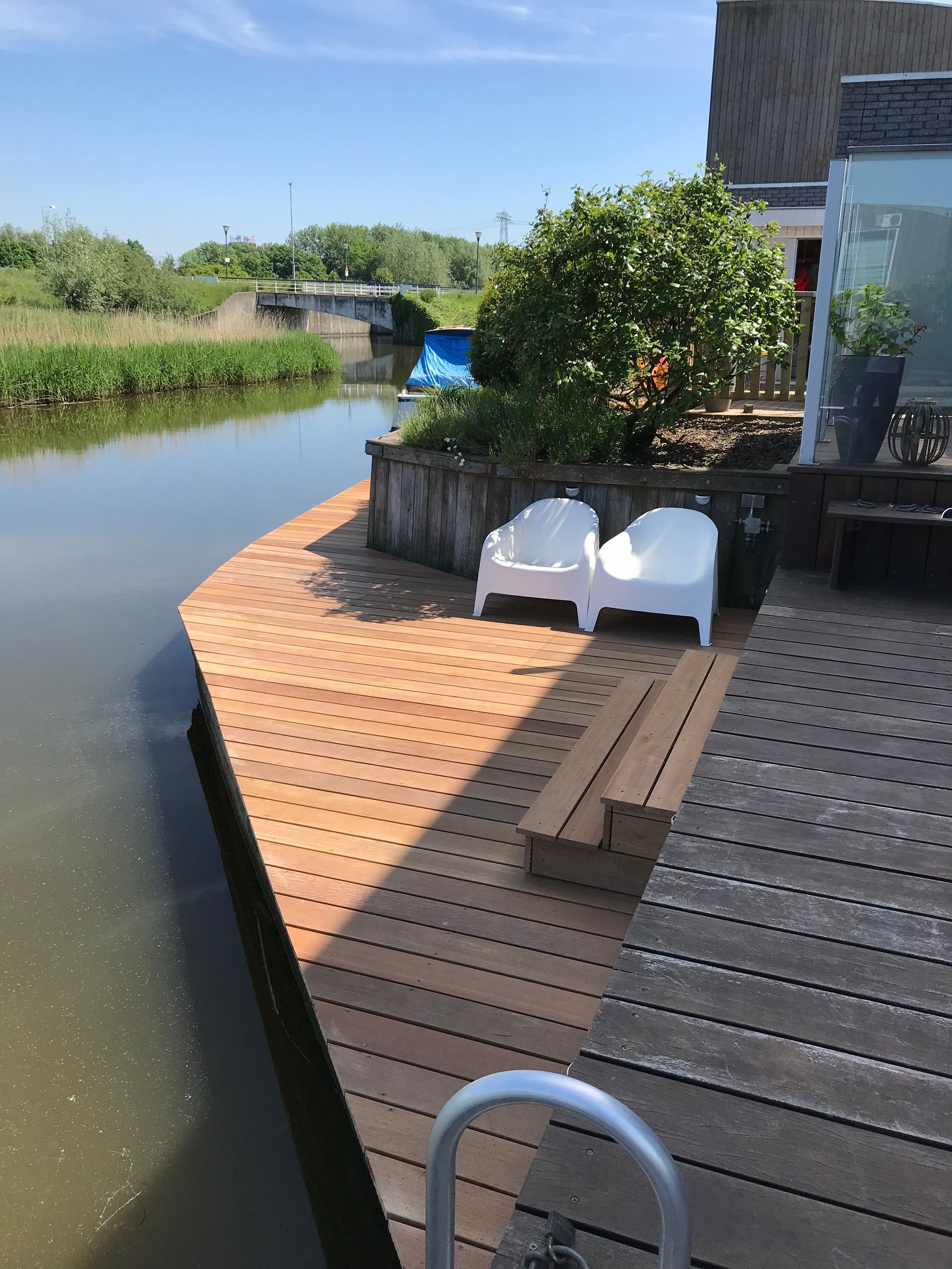 Een houten vlonder aan een kanaal met twee witte stoelen, een trap en wat groen, met uitzicht op een brug onder een helderblauwe hemel.
