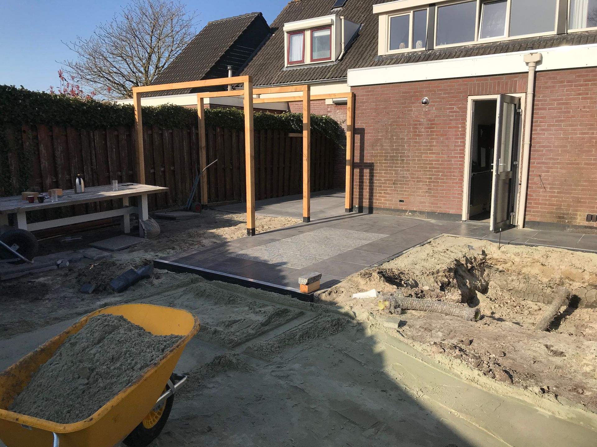 Een achtertuinrenovatie in uitvoering met een houten pergolaframe, stenen bestrating en een gele kruiwagen gevuld met grind.