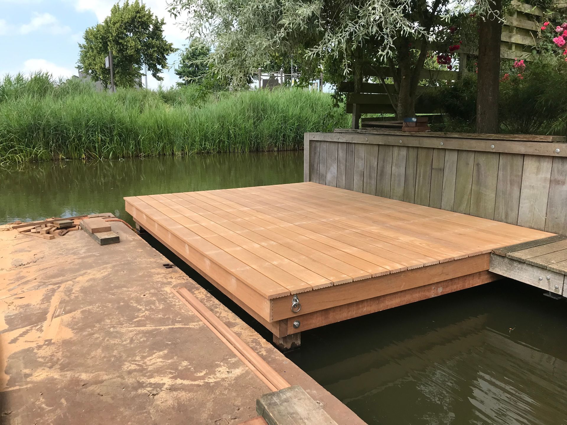 Een nieuwe, lichtgekleurde houten drijvende steiger ligt op kalm water naast een verweerde betonnen oever en weelderig groen.