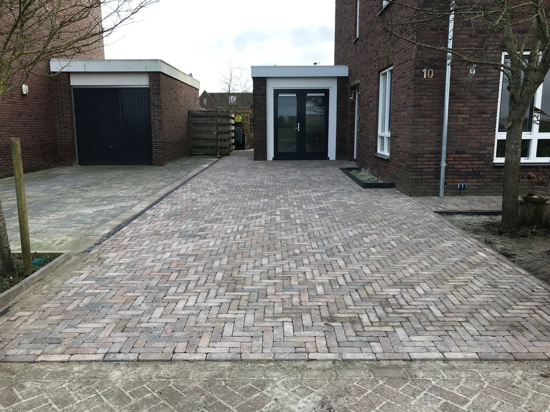 Een brede oprit, geplaveid met veelkleurige rechthoekige bakstenen, leidt naar de ingang van een bakstenen huis met een garage.
