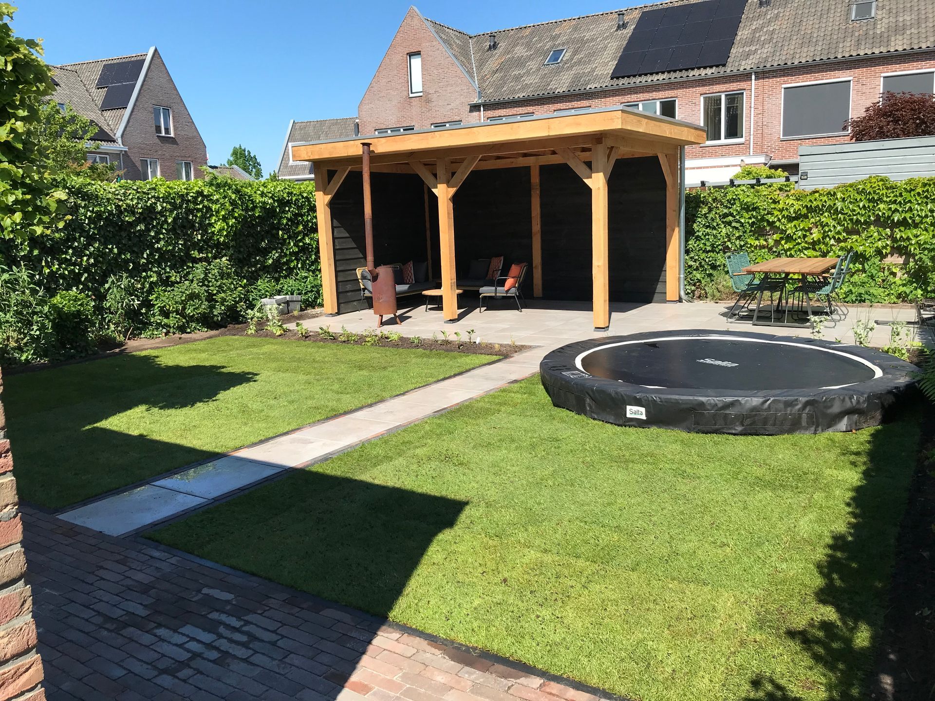 Een achtertuin met een groen gazon, een stenen pad, een ingebouwde trampoline en een houten prieel met zitgelegenheid buiten.