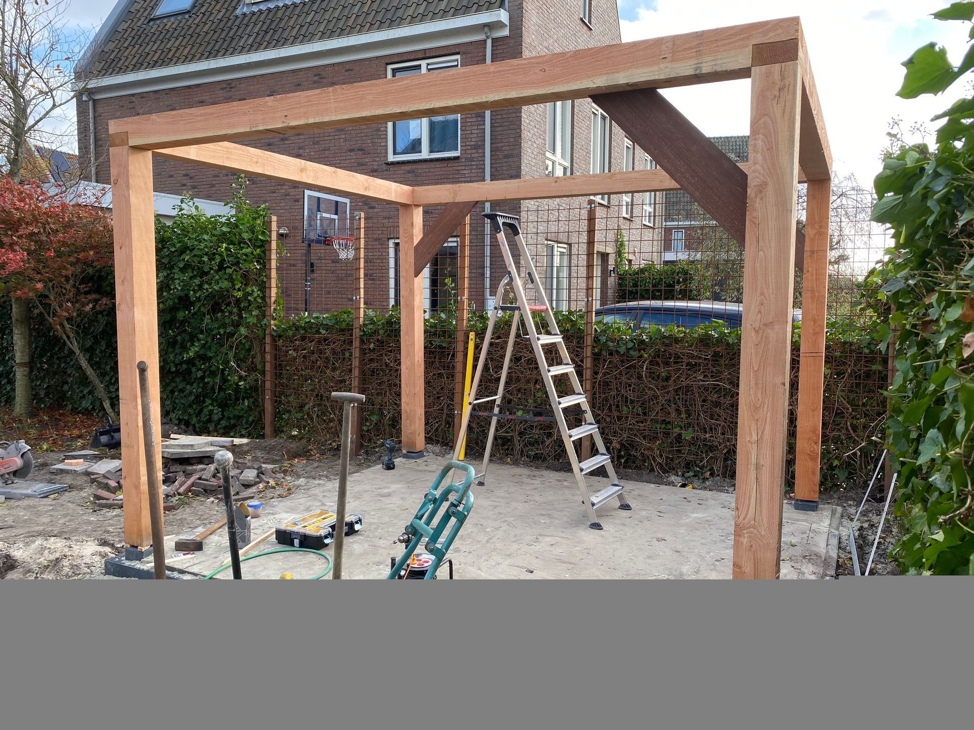 In een achtertuin, met een huis, een heg en een ladder zichtbaar, staat een gedeeltelijk gebouwde houten pergola.