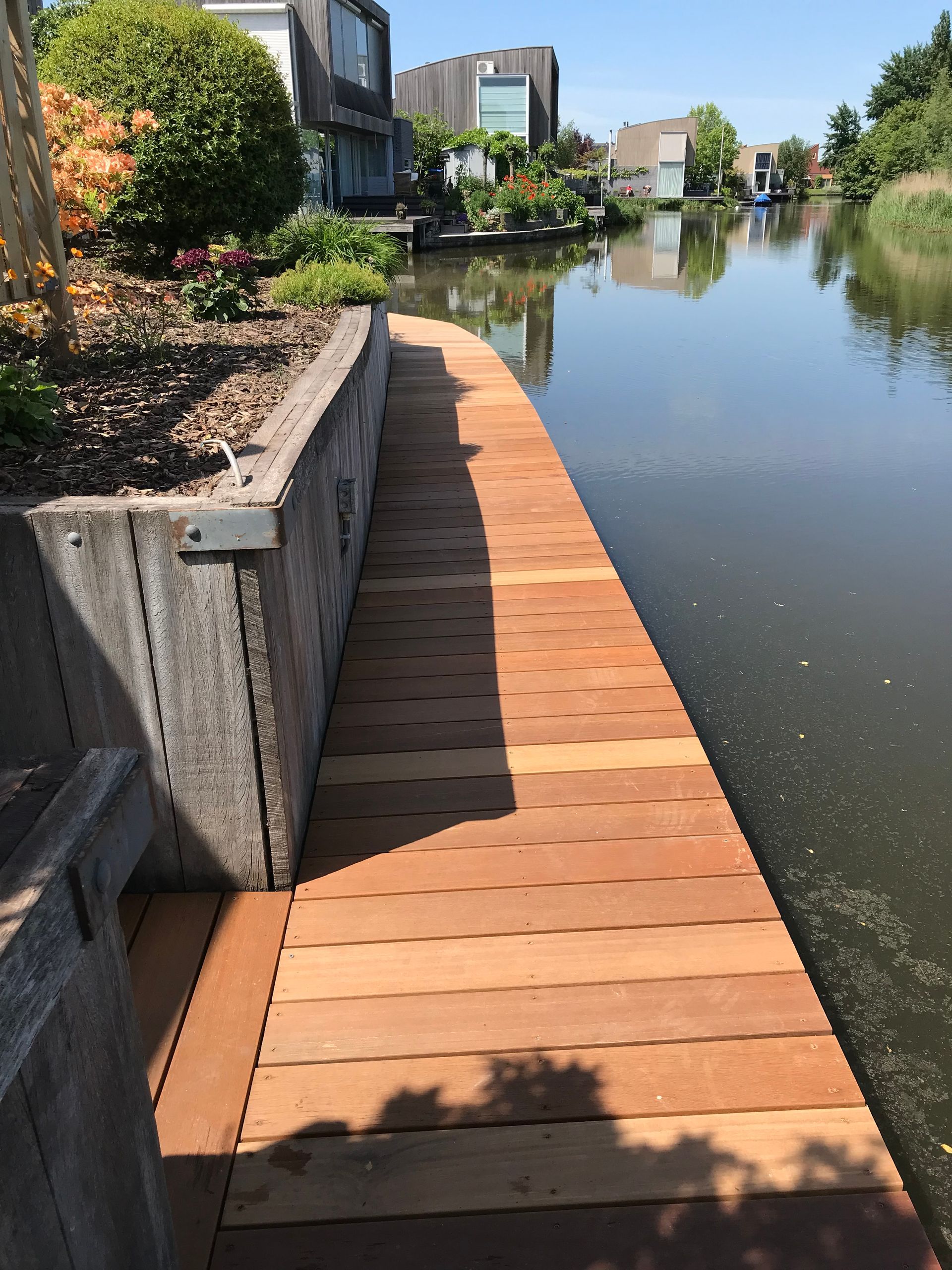 Een houten vlonderpad langs een kanaal, grenzend aan een bloembed met een stenen keermuur en moderne huizen op de achtergrond.