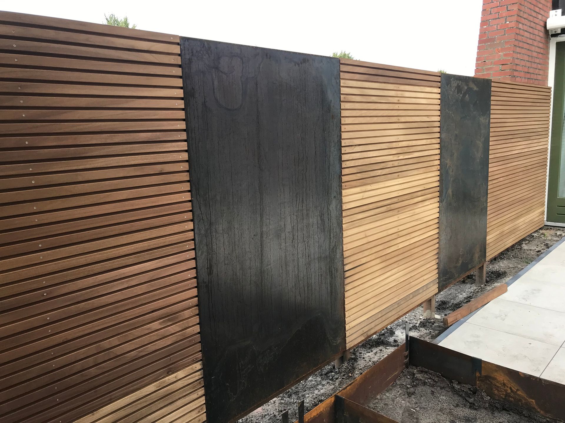 Een moderne tuinafscheiding met afwisselende panelen van horizontale houten latten en platte, verticale zwarte metalen platen.