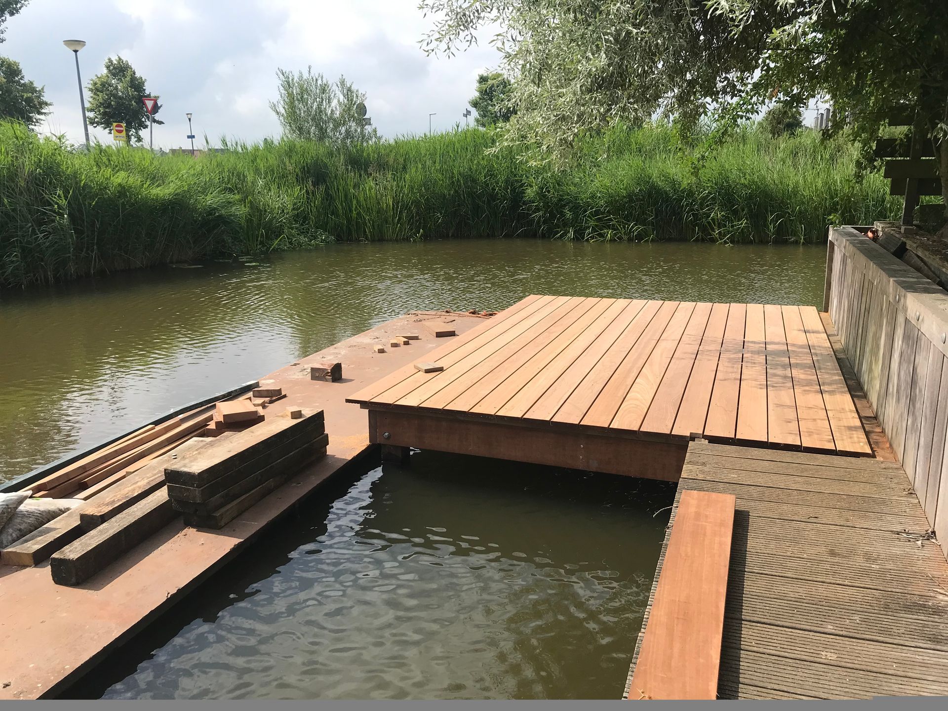 Een houten aanlegsteiger steekt uit boven het kalme water, naast een betonnen oever met structuur en weelderig groen riet.