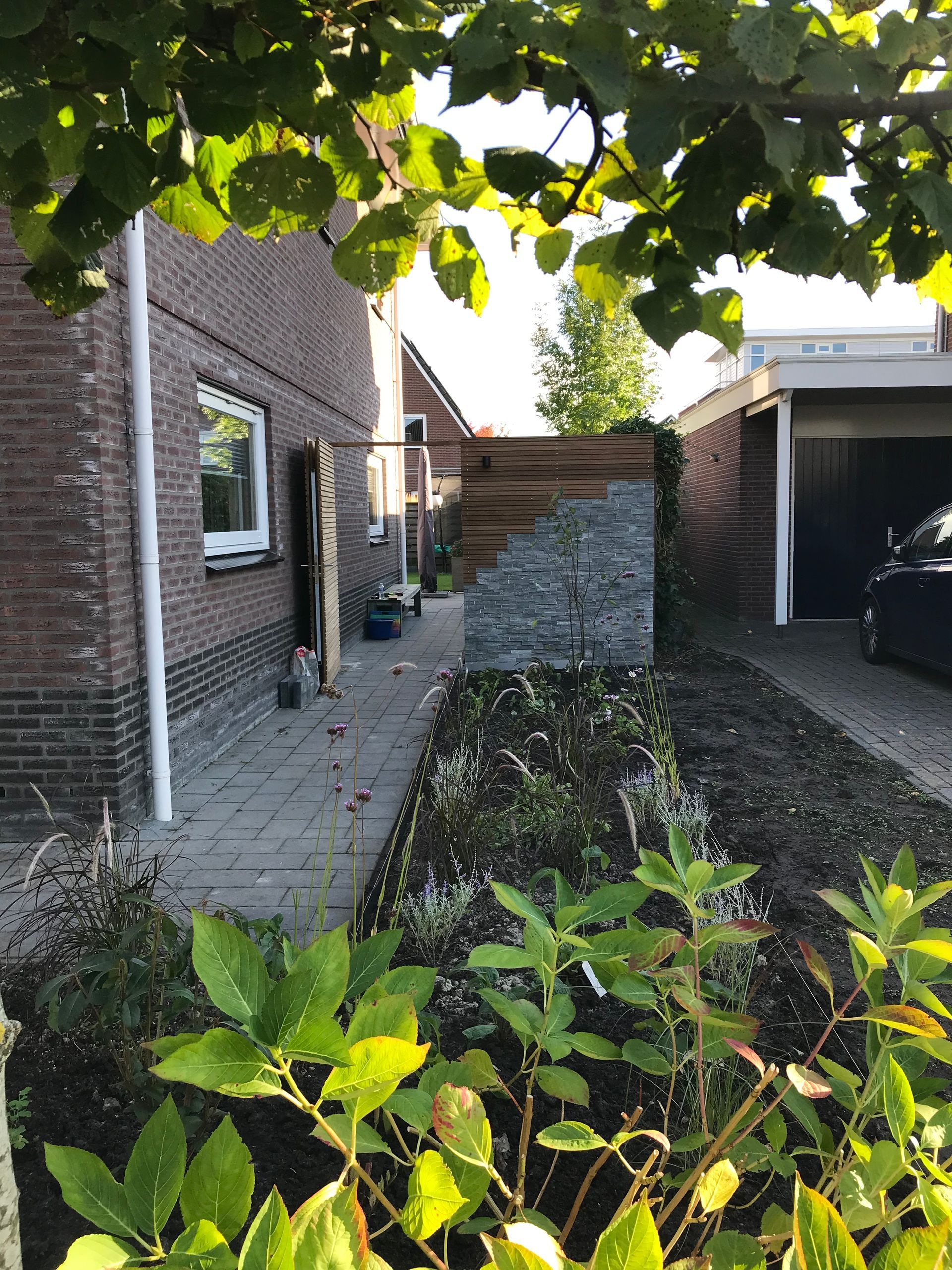 Zijaanzicht van een bakstenen huis met een bestraat pad, een stenen tuinafscheiding en een oprit ernaast, gezien door struiken heen.