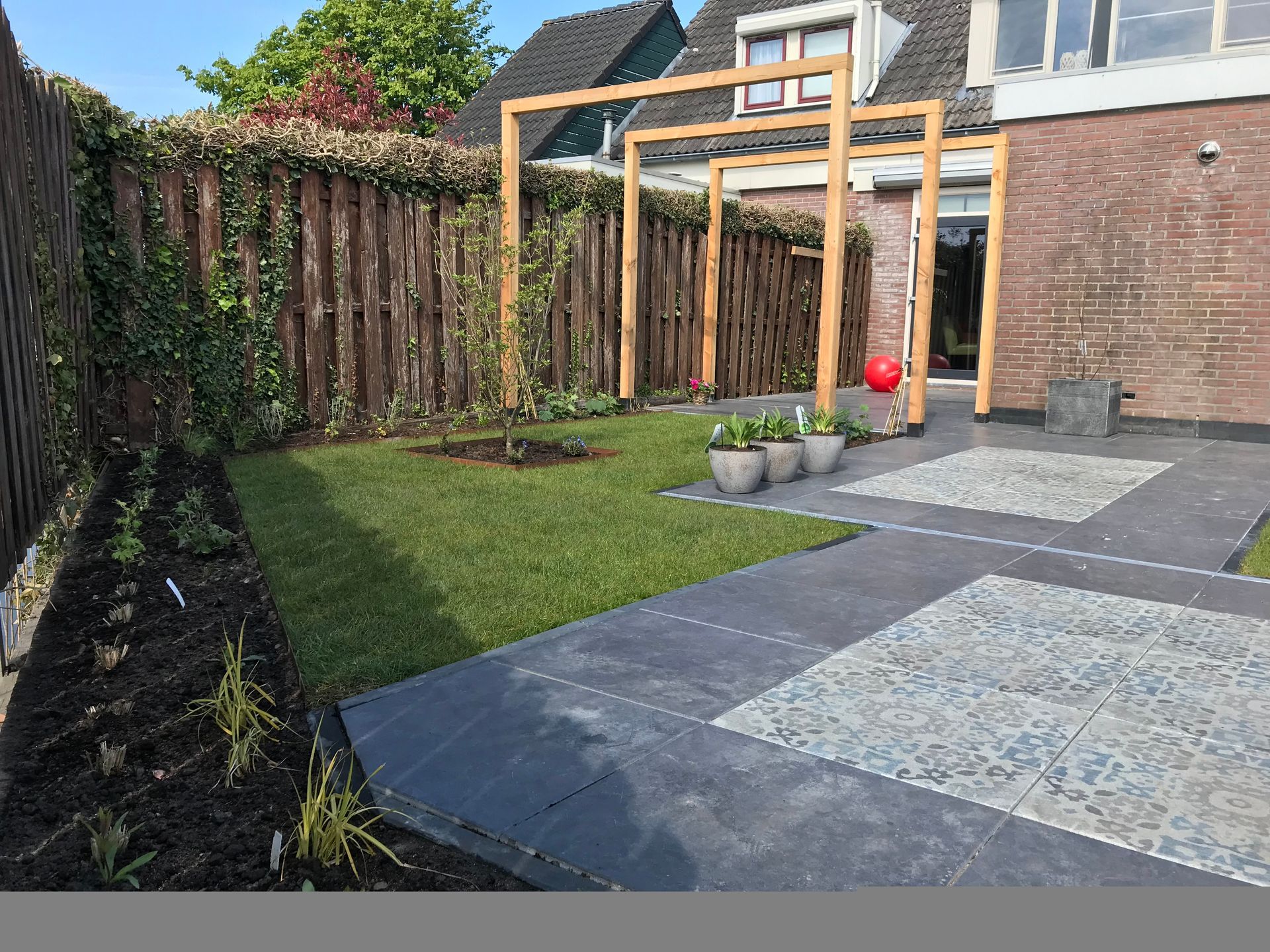 Een achtertuin met een gazon, een betegeld terras, een houten pergola en donkere mulch langs de schutting.