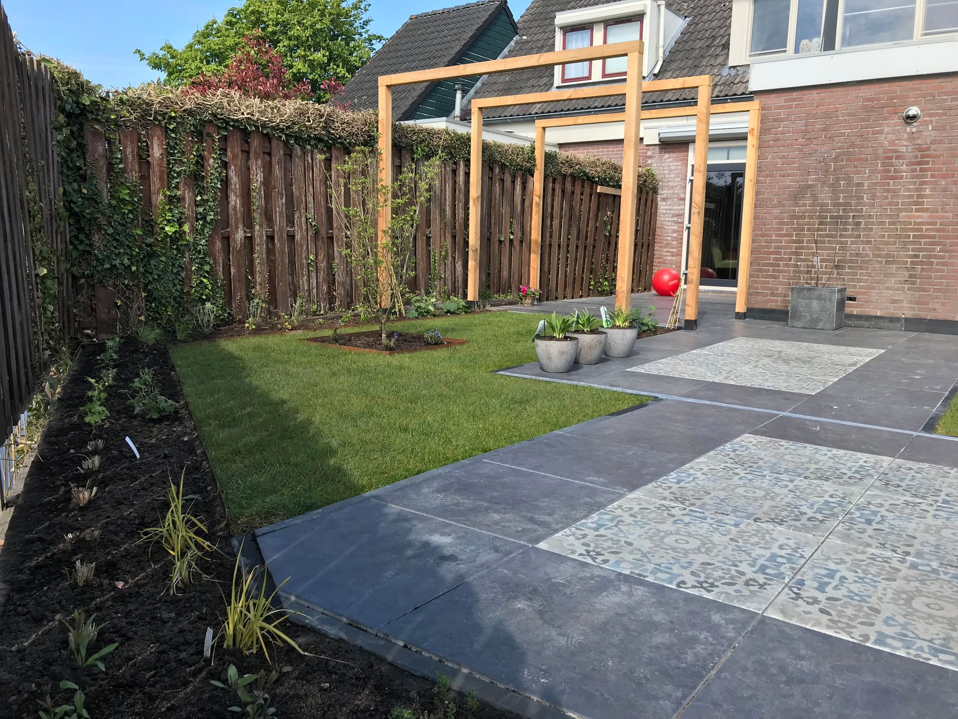Een moderne achtertuin met een gazon, een donker betegeld terras met stenen accenten, een houten pergola en een privacyscherm.