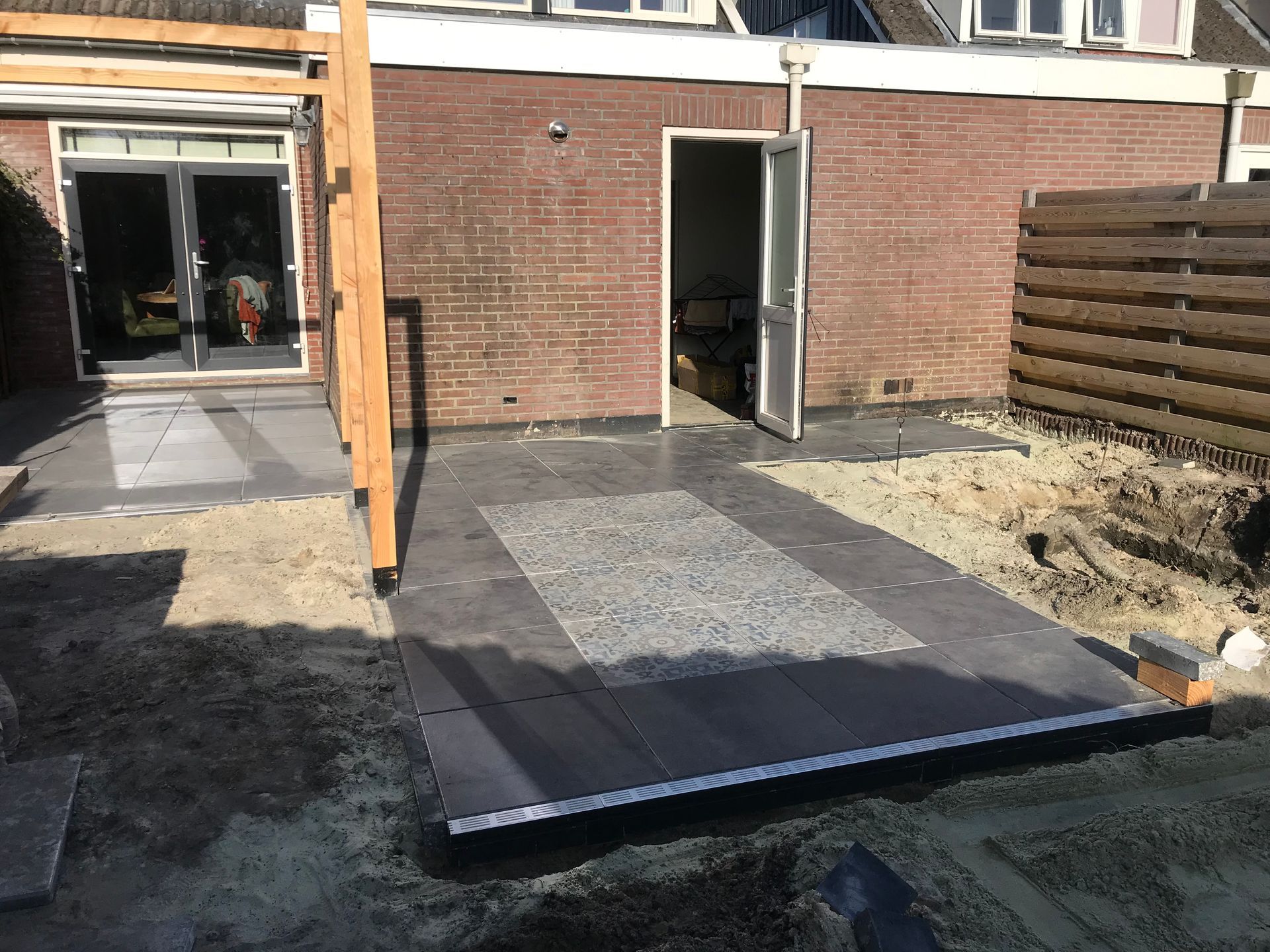 Een gedeeltelijk afgewerkt terras met donkere bestrating, een grindgedeelte in het midden en houten palen tegen een bakstenen huis.