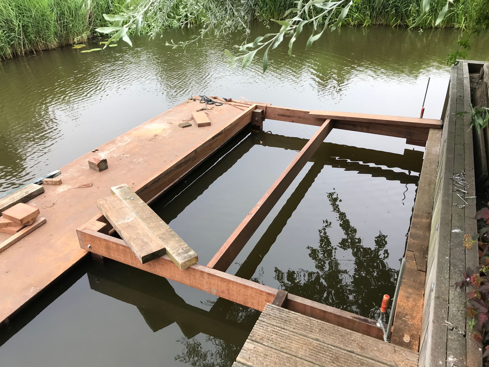 Een houten terras in aanbouw steekt uit boven een kalm wateroppervlak, waarbij een deel van het frame zichtbaar is.