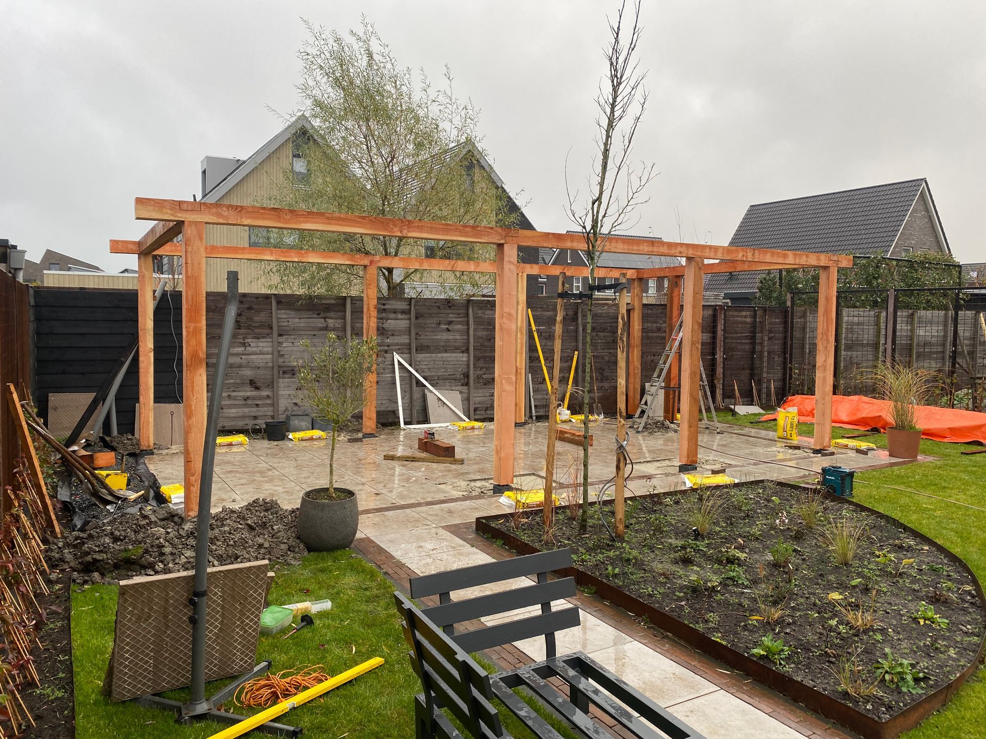 Een houten pergolaframe in aanbouw in een achtertuin in een buitenwijk, met een betegeld terras en een aangelegde bloembed.