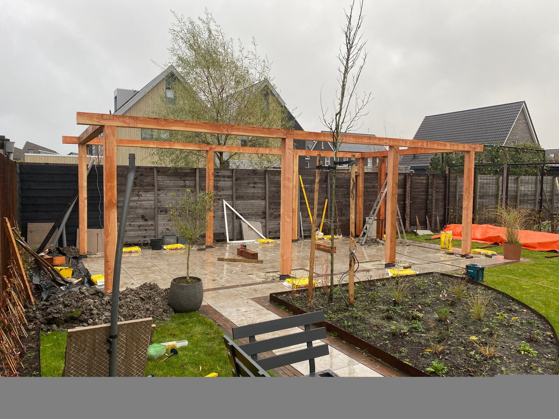 Een houten pergolaframe staat boven een terras in een achtertuin die in aanbouw is, met een gazon en bloembedden in de buurt.