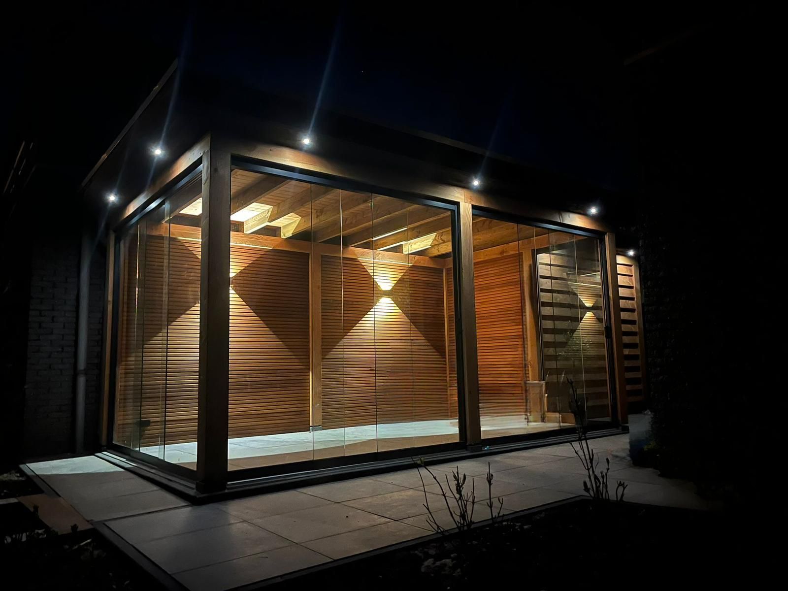 Een moderne, houten tuinconstructie met glazen wanden en warme, sfeervolle verlichting 's nachts.