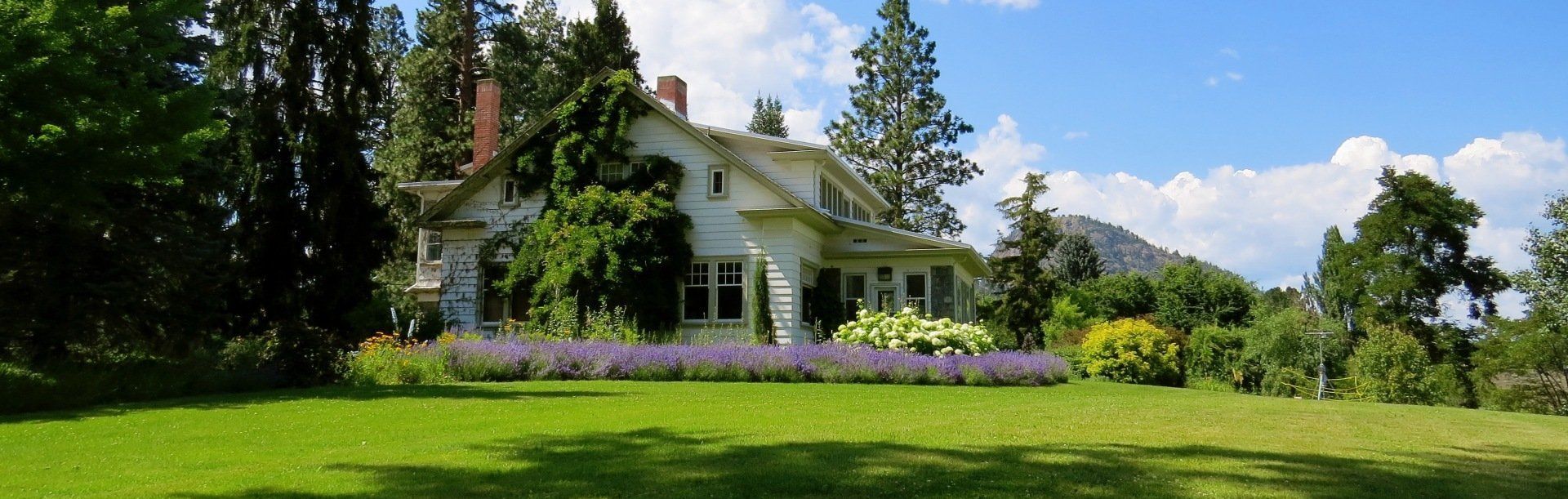 Een wit, twee verdiepingen tellend huis met een groen gazon en paarse bloemen staat tussen de bomen onder een blauwe hemel met zachte wolken.