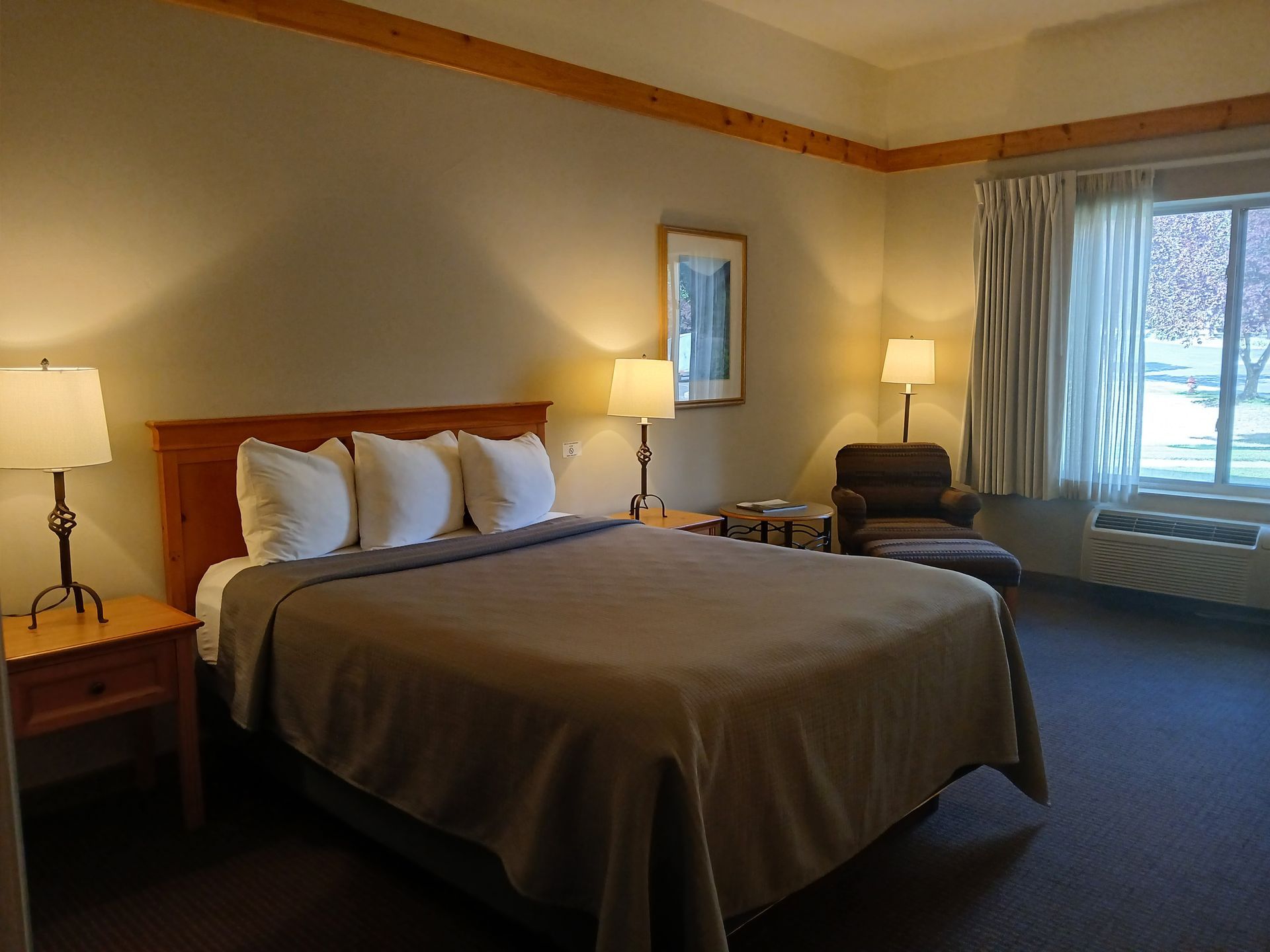 A hotel room with a king size bed , desk , chair and lamps.