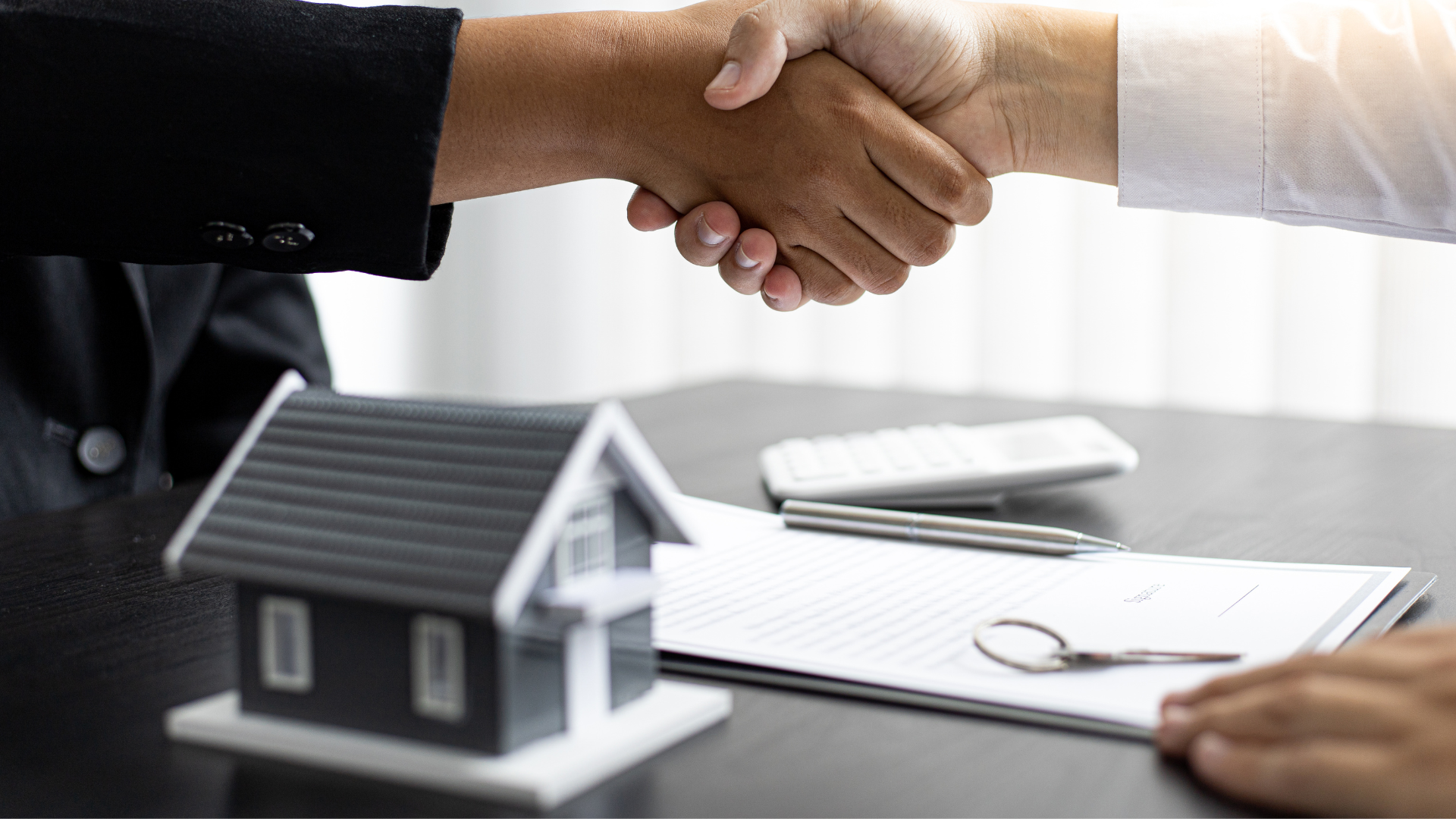 Handshake over desk beside house model and paperwork, suggesting a real estate deal