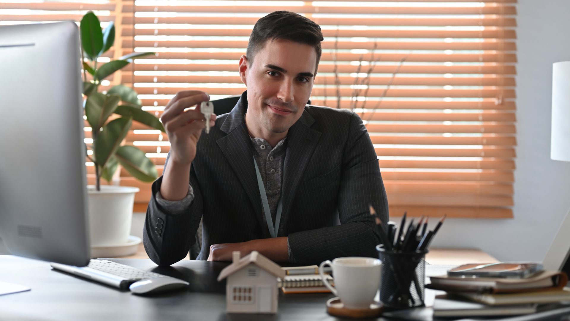 Man at a desk in an office, holding a pen and looking toward the camera, with blinds and plants behind him.