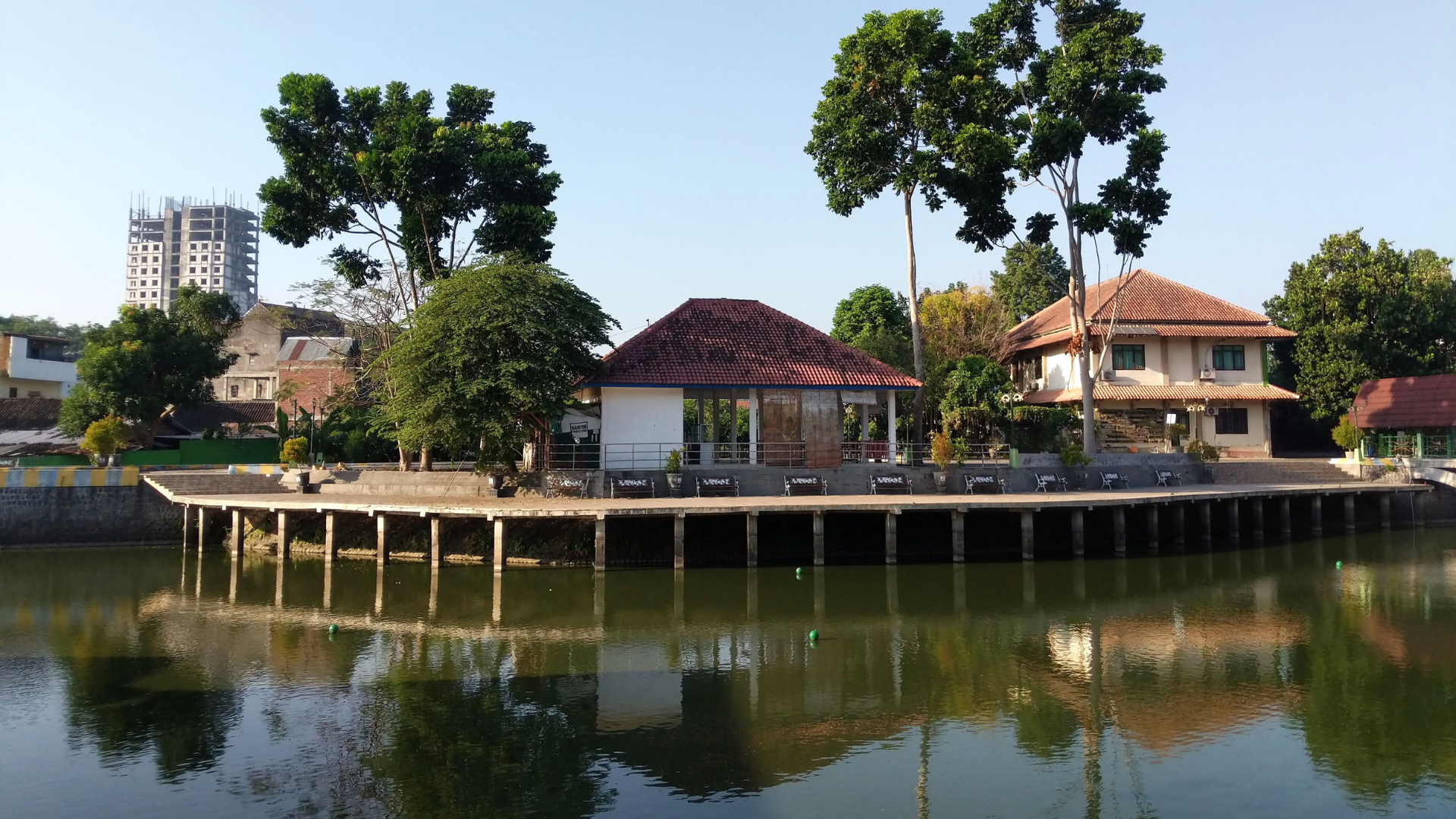 Black house with a red roof on a lake, surrounded by green trees.