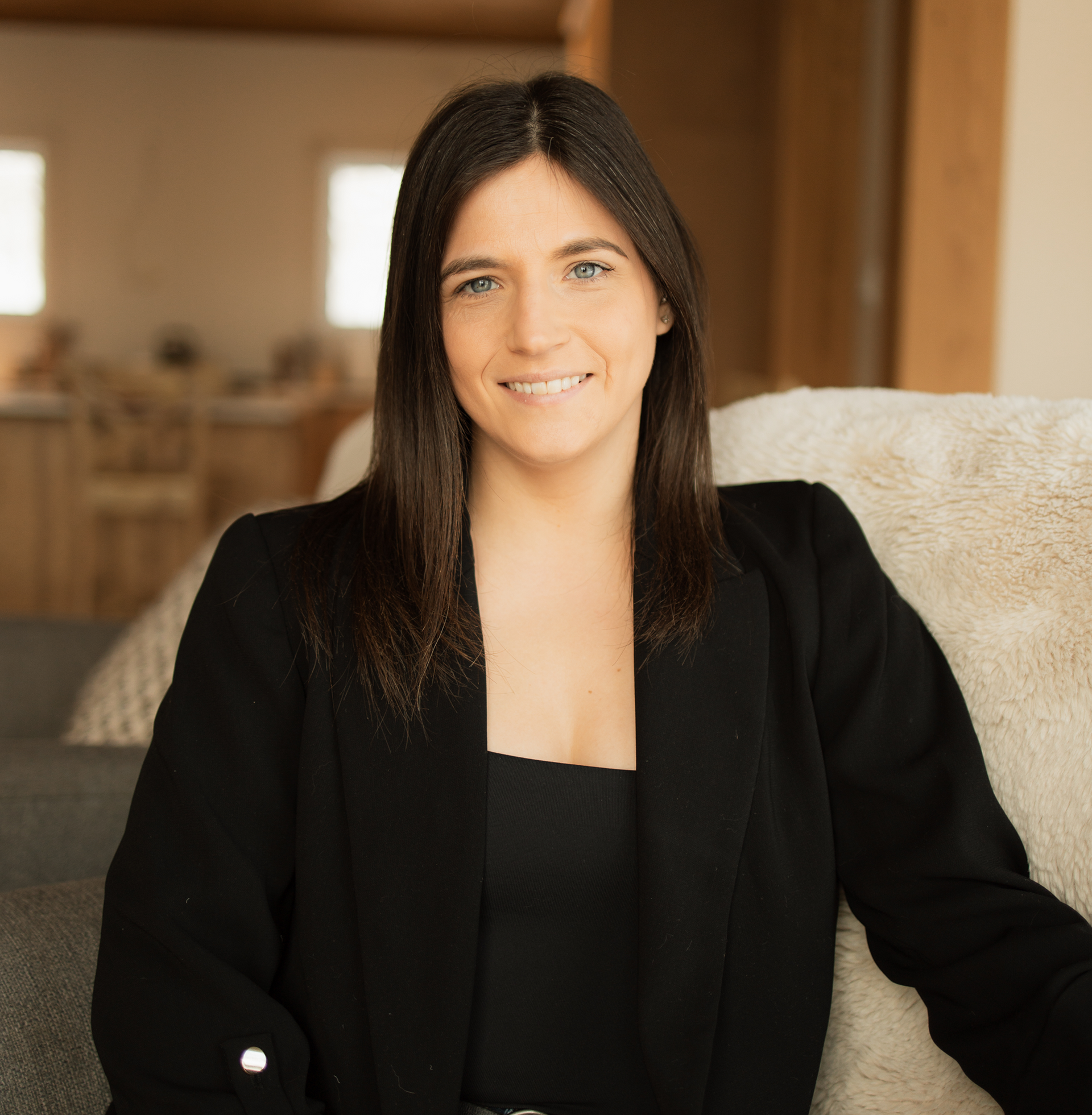 A woman in a black jacket is smiling while sitting on a couch.