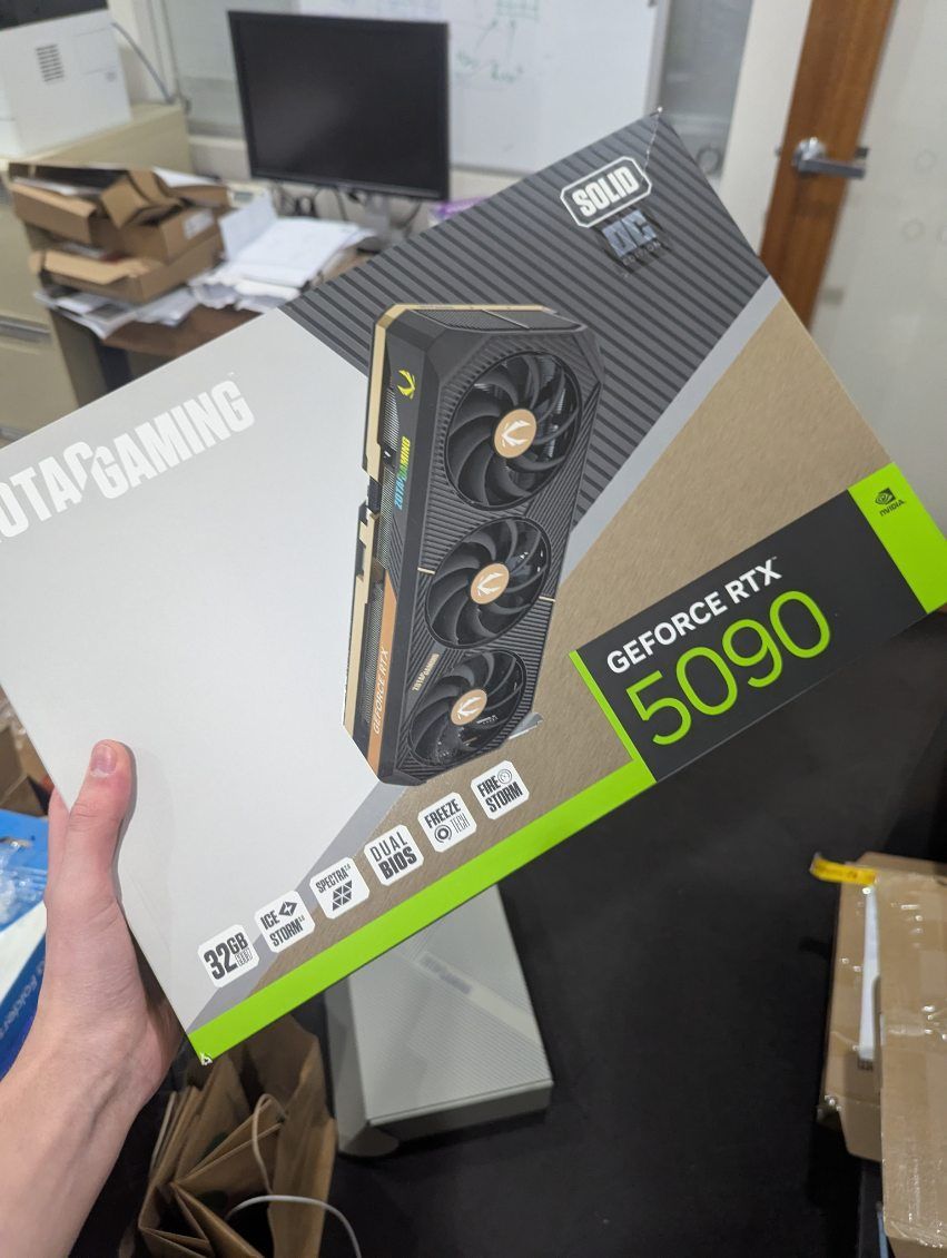 Person holding an MSI GeForce RTX 5090 graphics card box in a cluttered room.