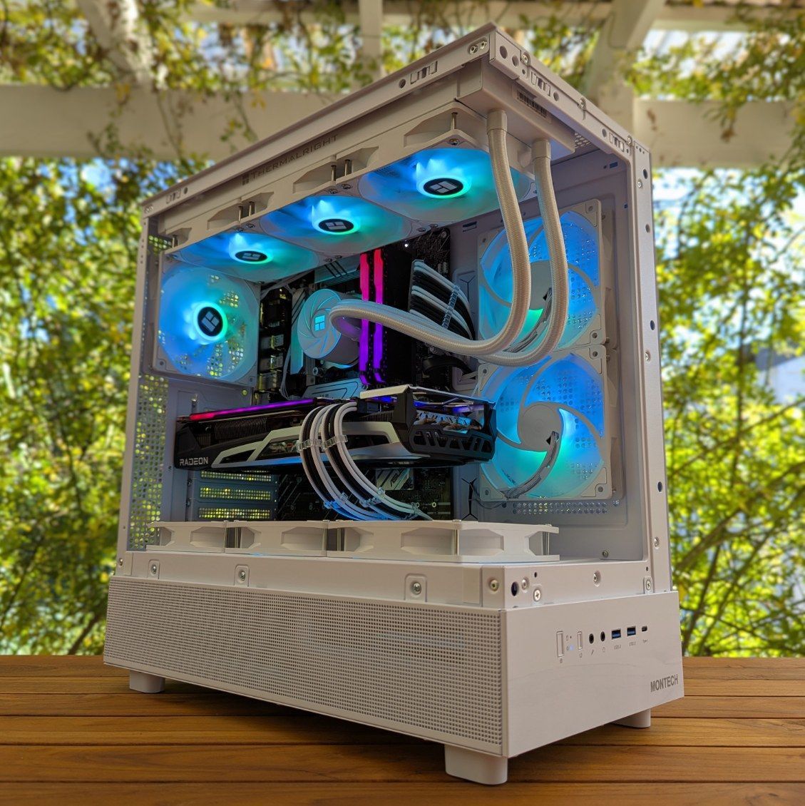 White custom desktop PC with blue RGB-lit fans and water cooling on a wooden table outdoors