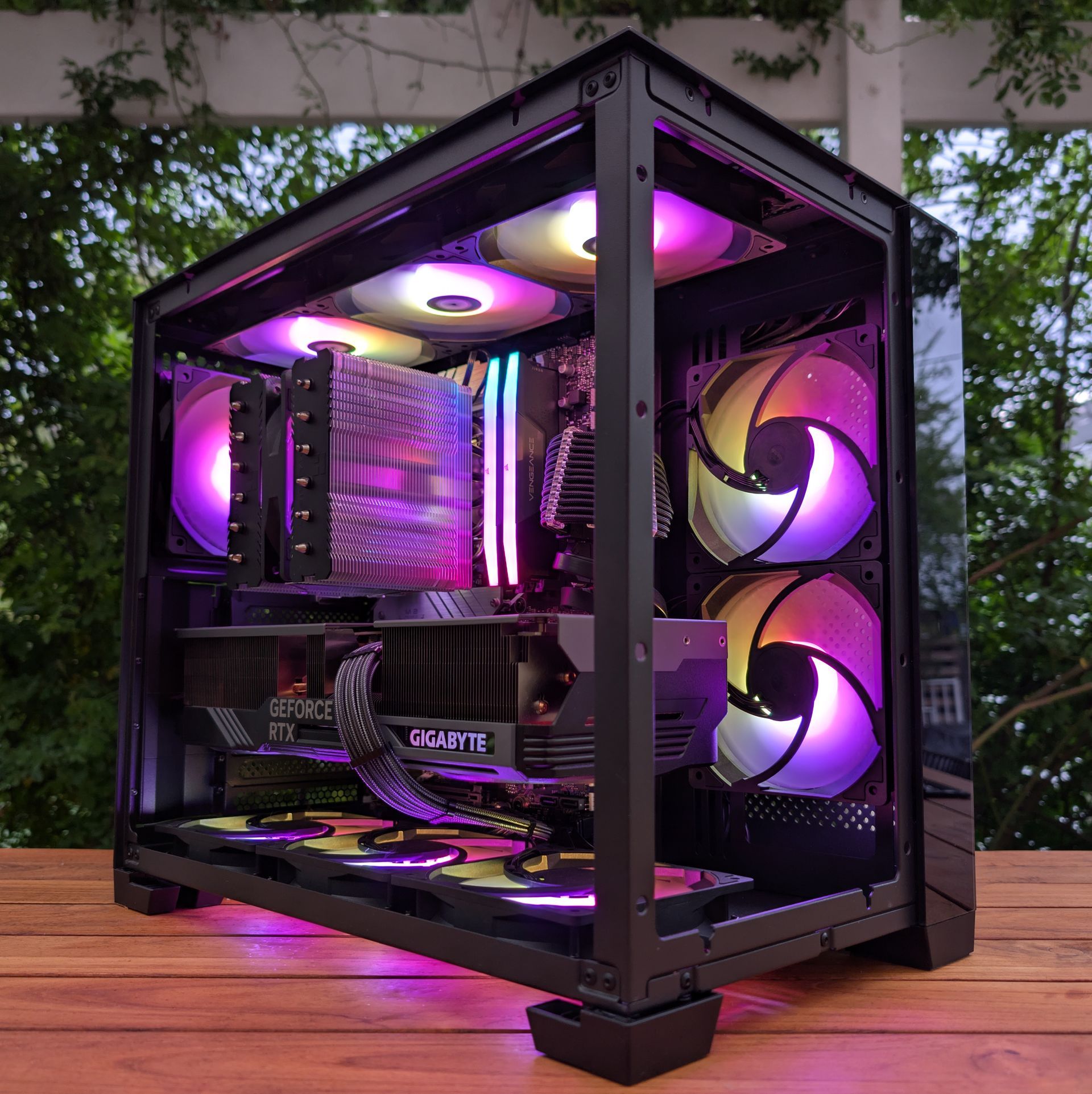 RGB-lit custom gaming PC in a glass case on a wooden table outdoors, with purple fans and lighting