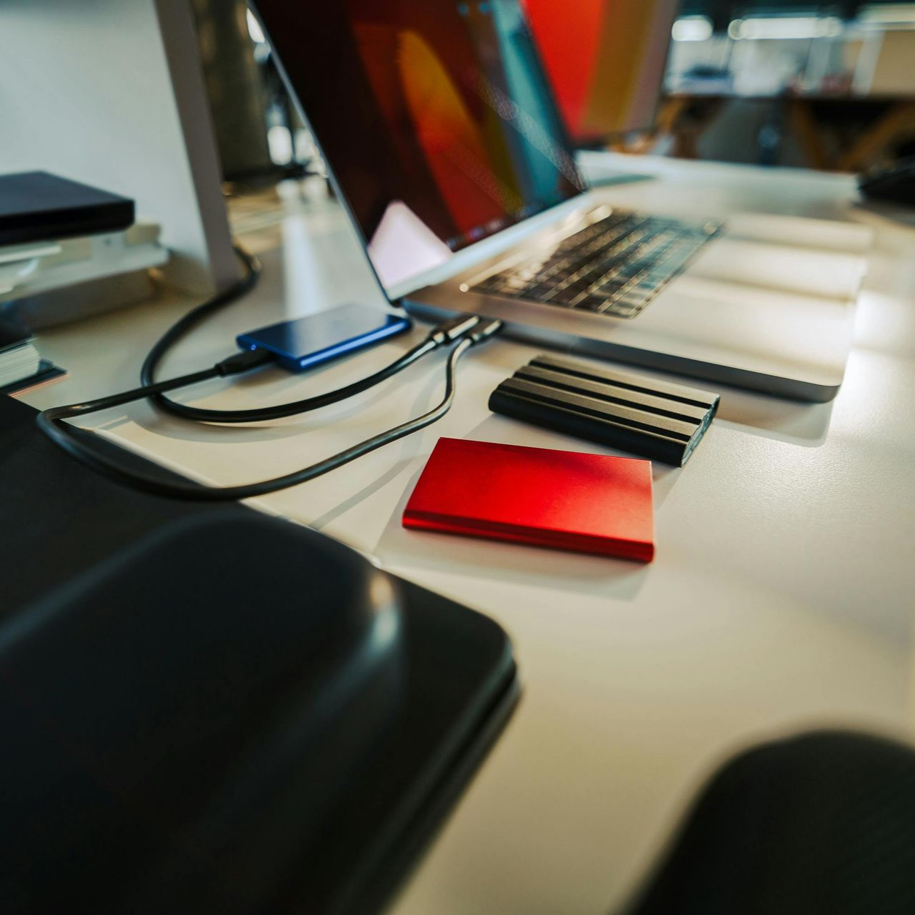 Laptop on a desk with cables, a blue device, black notebook, and red card in a bright office setting
