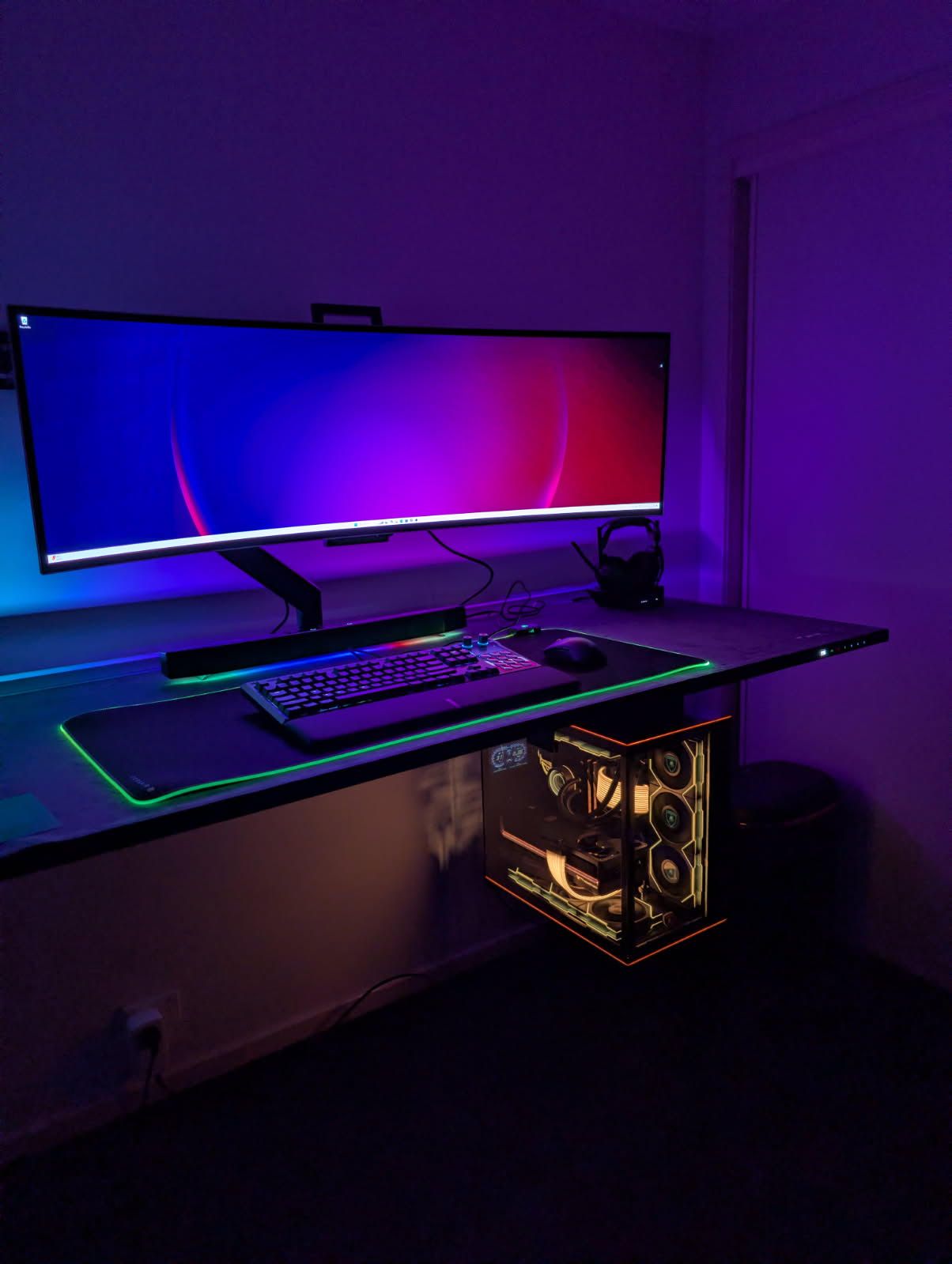 RGB-lit gaming desk with ultrawide monitor, keyboard, and glass PC tower in a dark room