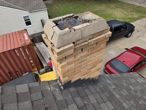 Damaged brick chimney on a shingled roof beside parked red and black cars, with debris at the top