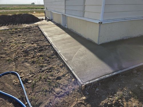 Concrete walkway beside a house foundation beside bare dirt and a garden hose in a yard.